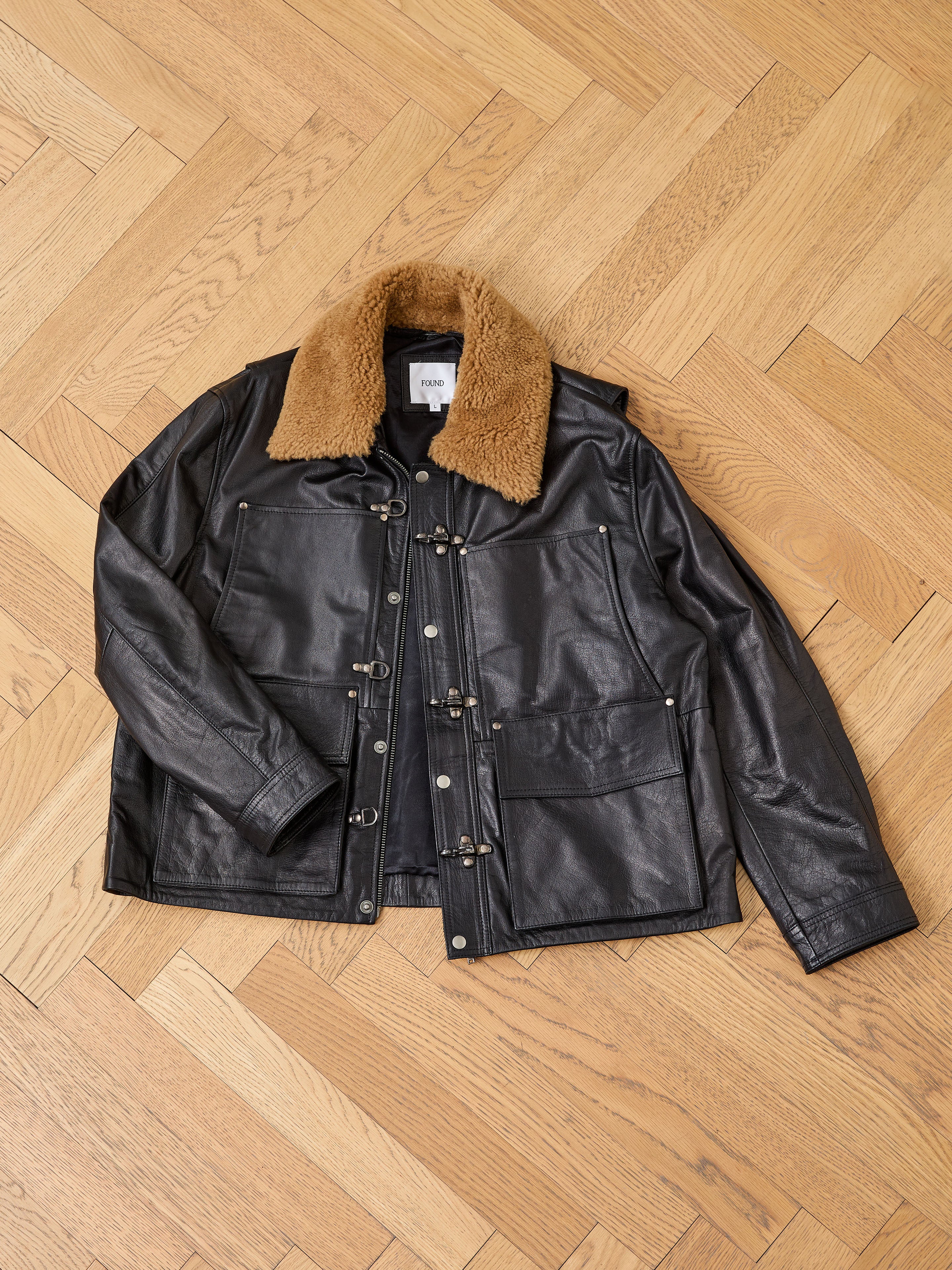 A FOUND Sherpa Collar Leather Utility Jacket in black, made from genuine leather with large front pockets and a brown sherpa collar, is laid flat on a wooden parquet floor.