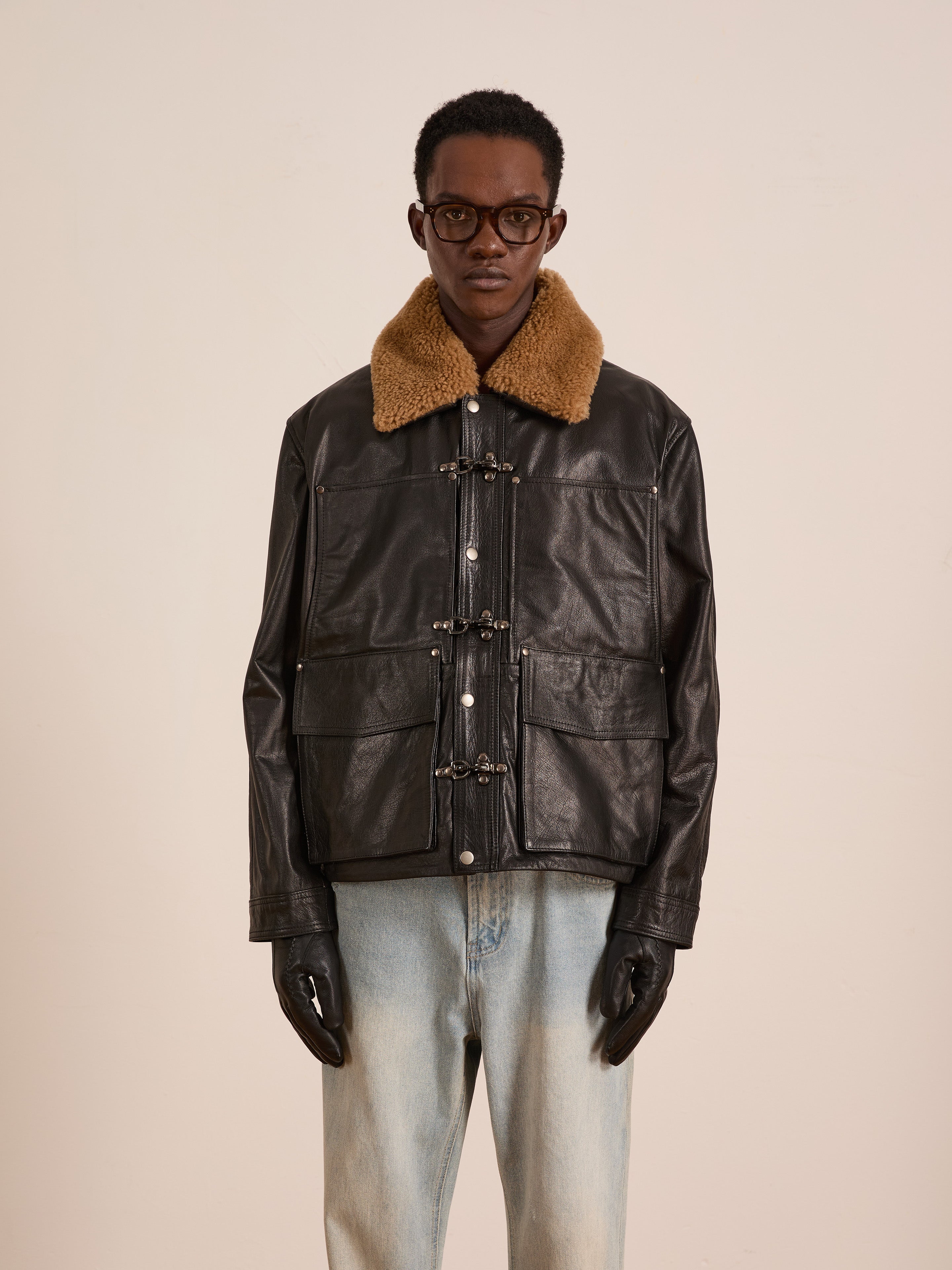 A person wearing the FOUND Sherpa Collar Leather Utility Jacket, light blue jeans, black gloves, and glasses stands against a plain light background.