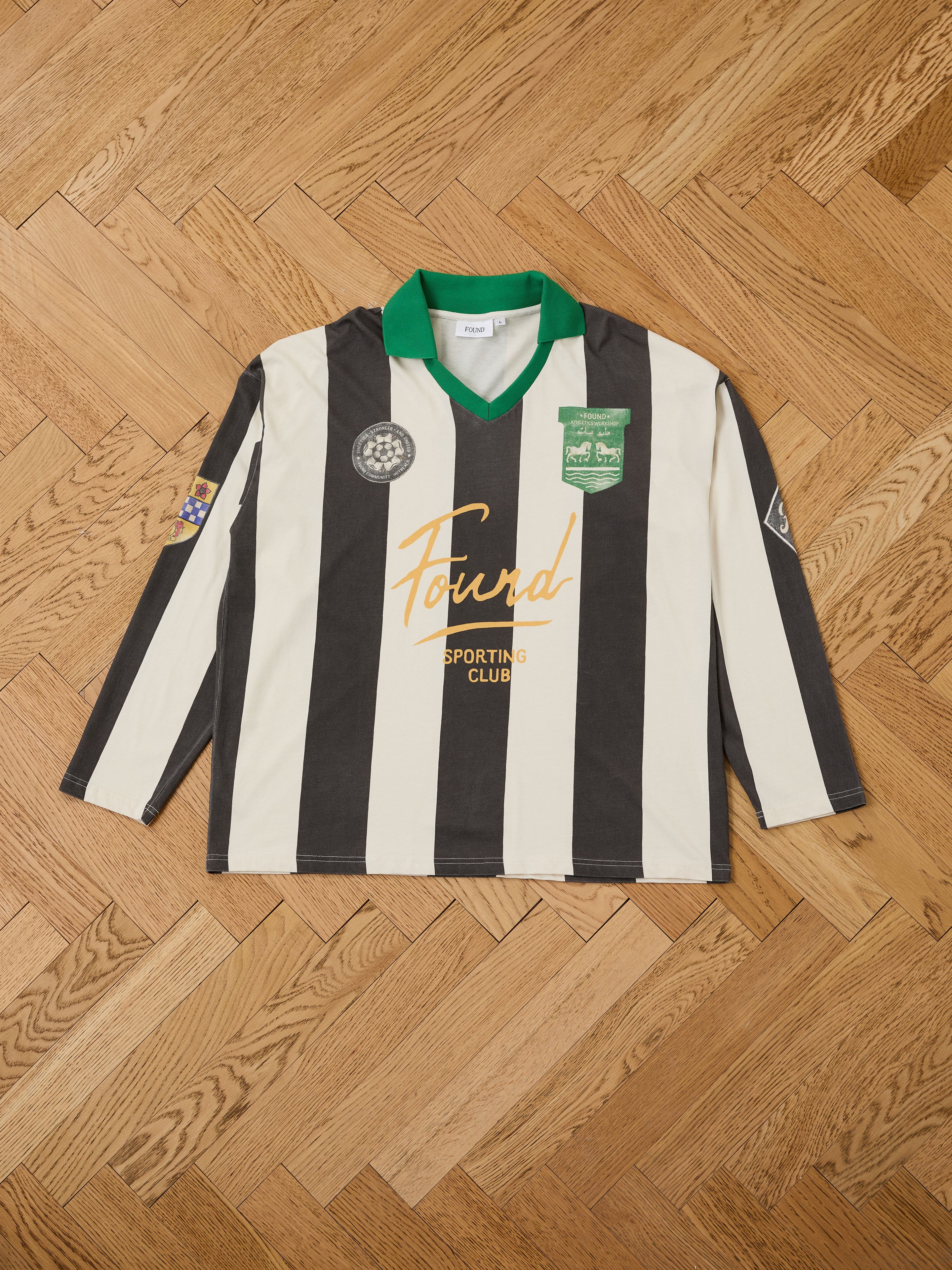 The FOUND Striped Sport LS Jersey features a relaxed unisex fit, black and white stripes, green collar, gold "Found Sporting Club" text, and multiple patches. Shown laid flat on a wooden floor.