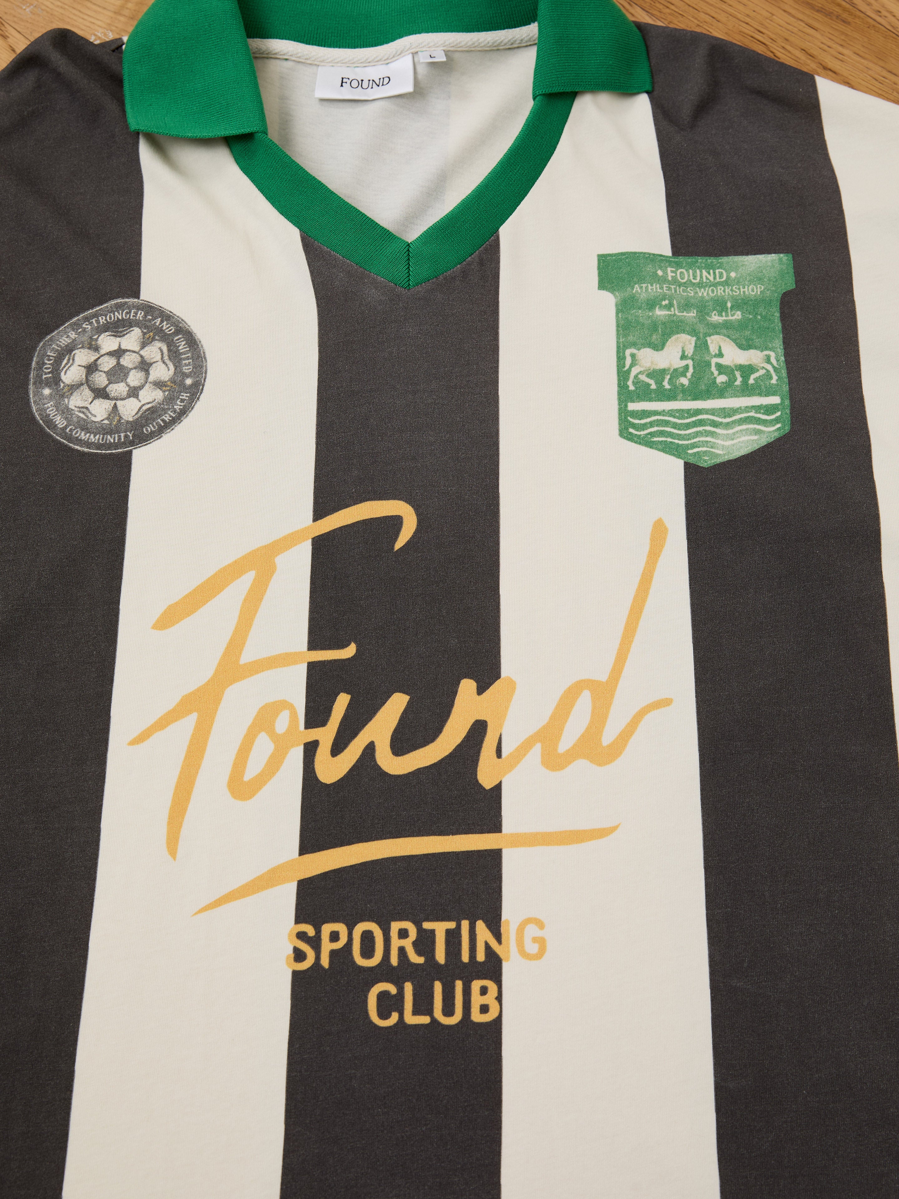 The FOUND Striped Sport LS Jersey features black and white stripes, a green collar, relaxed unisex fit, two patches, and "Found Sporting Club" in yellow on the front for versatile style.