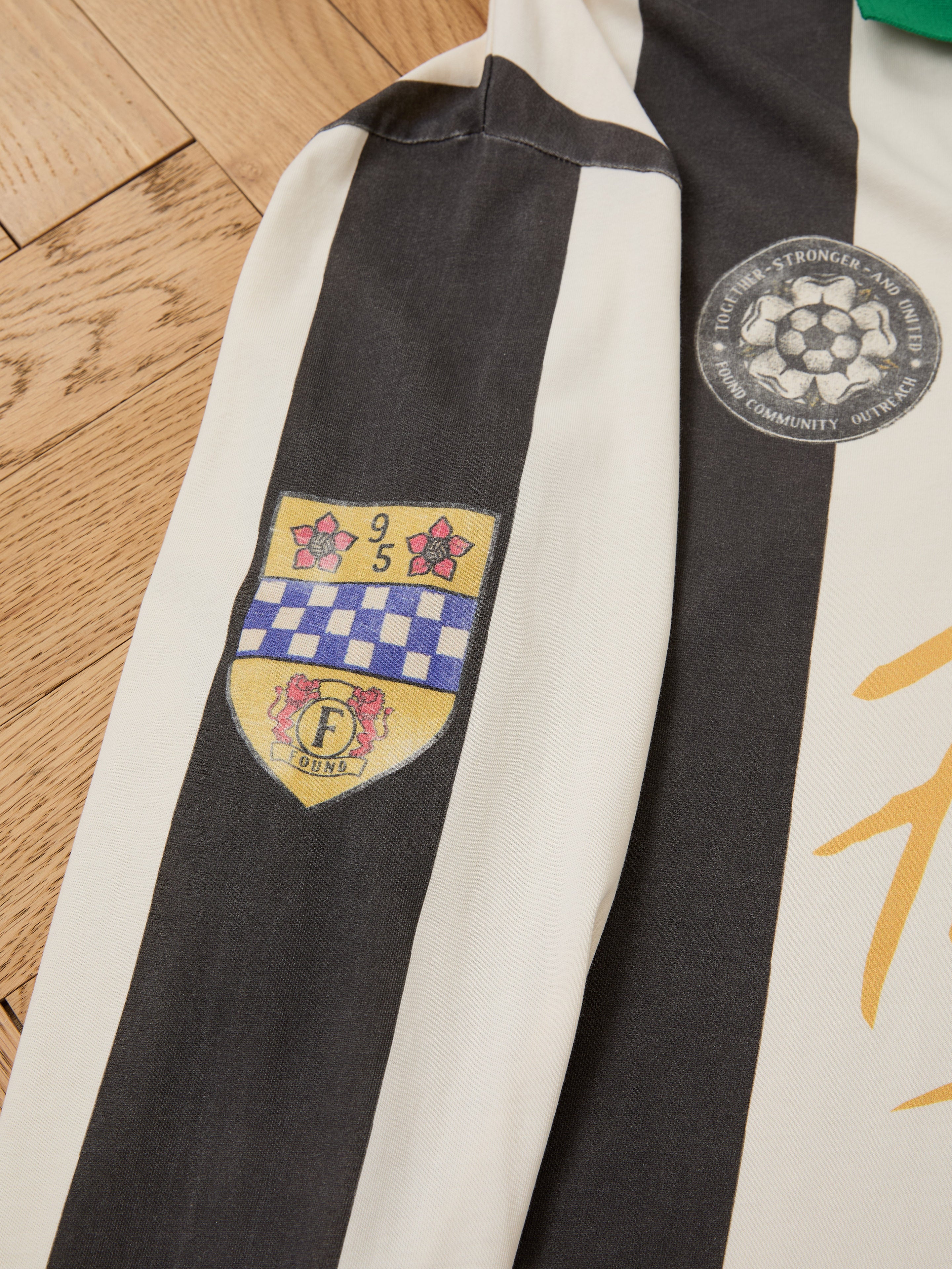 A close-up of the FOUND Striped Sport LS Jersey sleeve, unisex relaxed fit, shows two embroidered patches—a shield and a football silhouette—on a wooden floor.