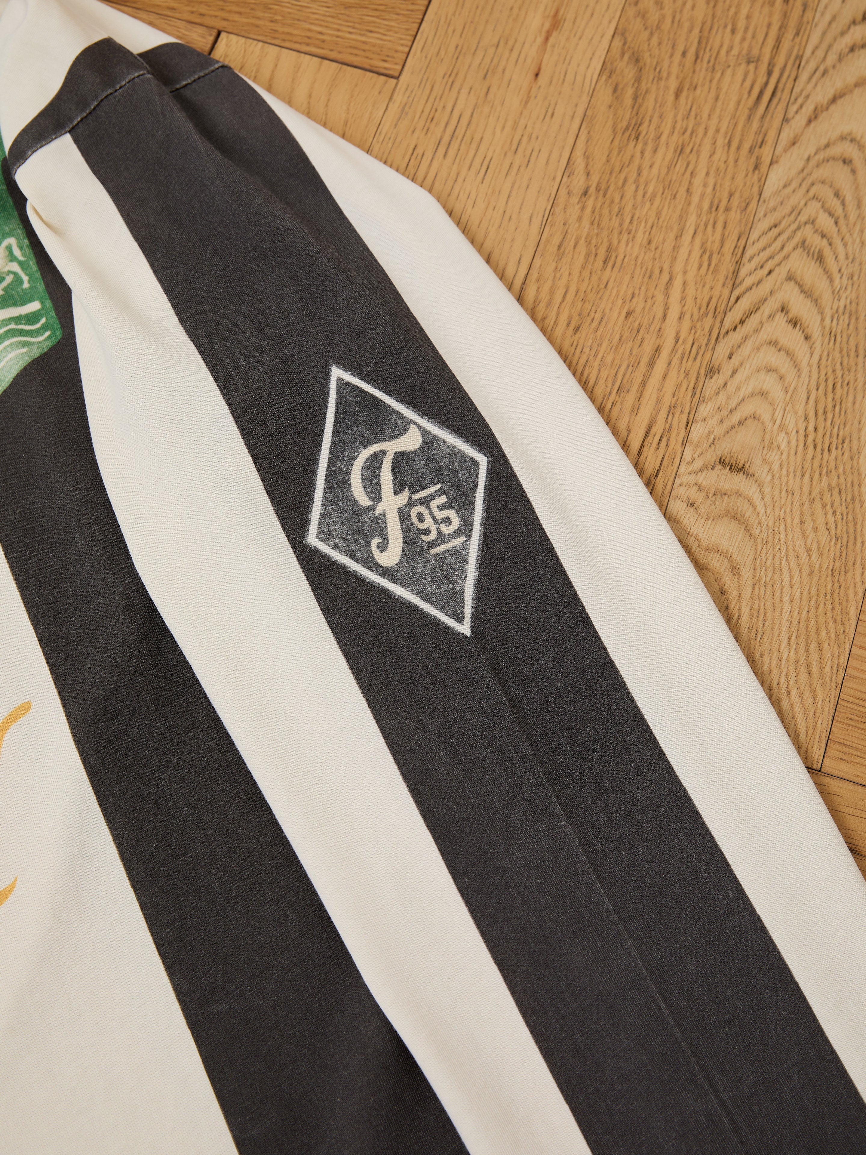 A close-up of the FOUND Striped Sport LS Jersey sleeve shows its black and white stripes with a diamond-shaped "F95" emblem, unisex relaxed fit, displayed on a wooden floor.