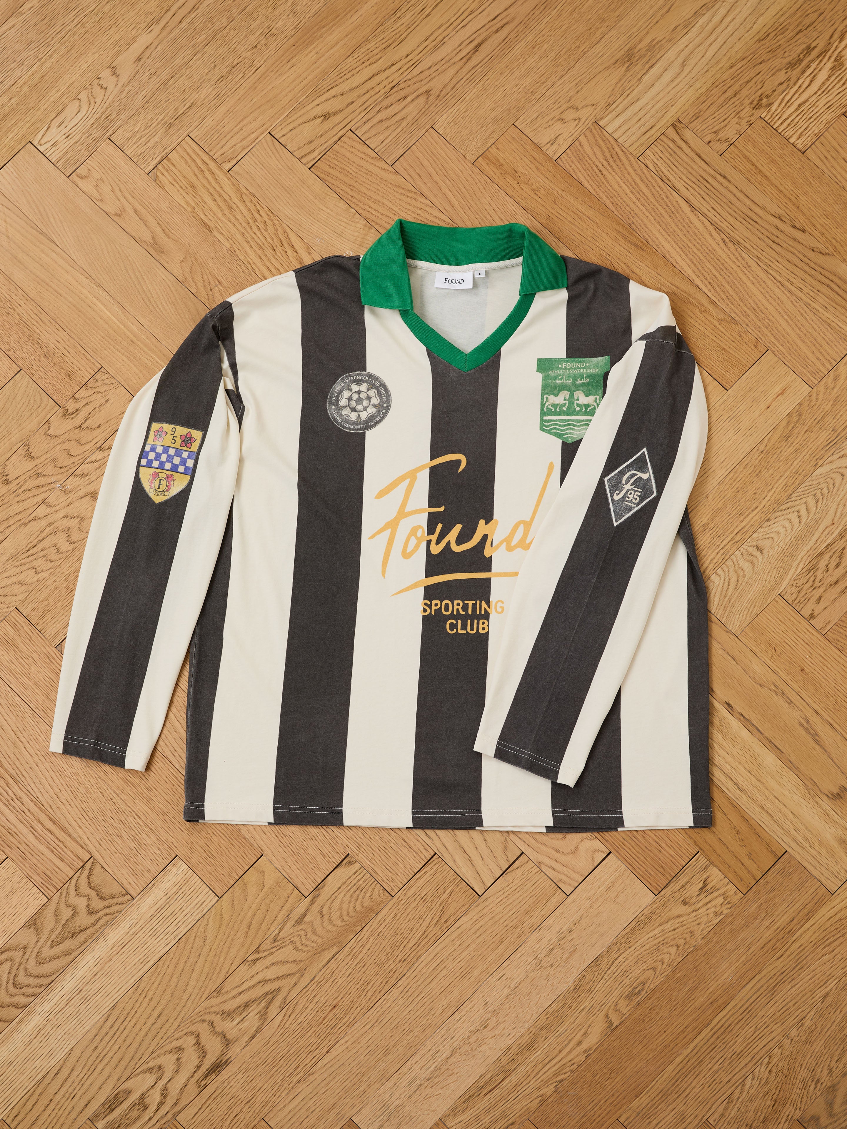 The FOUND Striped Sport LS Jersey is a unisex, relaxed fit long sleeve top with black and white stripes, green collar, sponsor logos, and sleeve patches, displayed flat on a wooden floor.