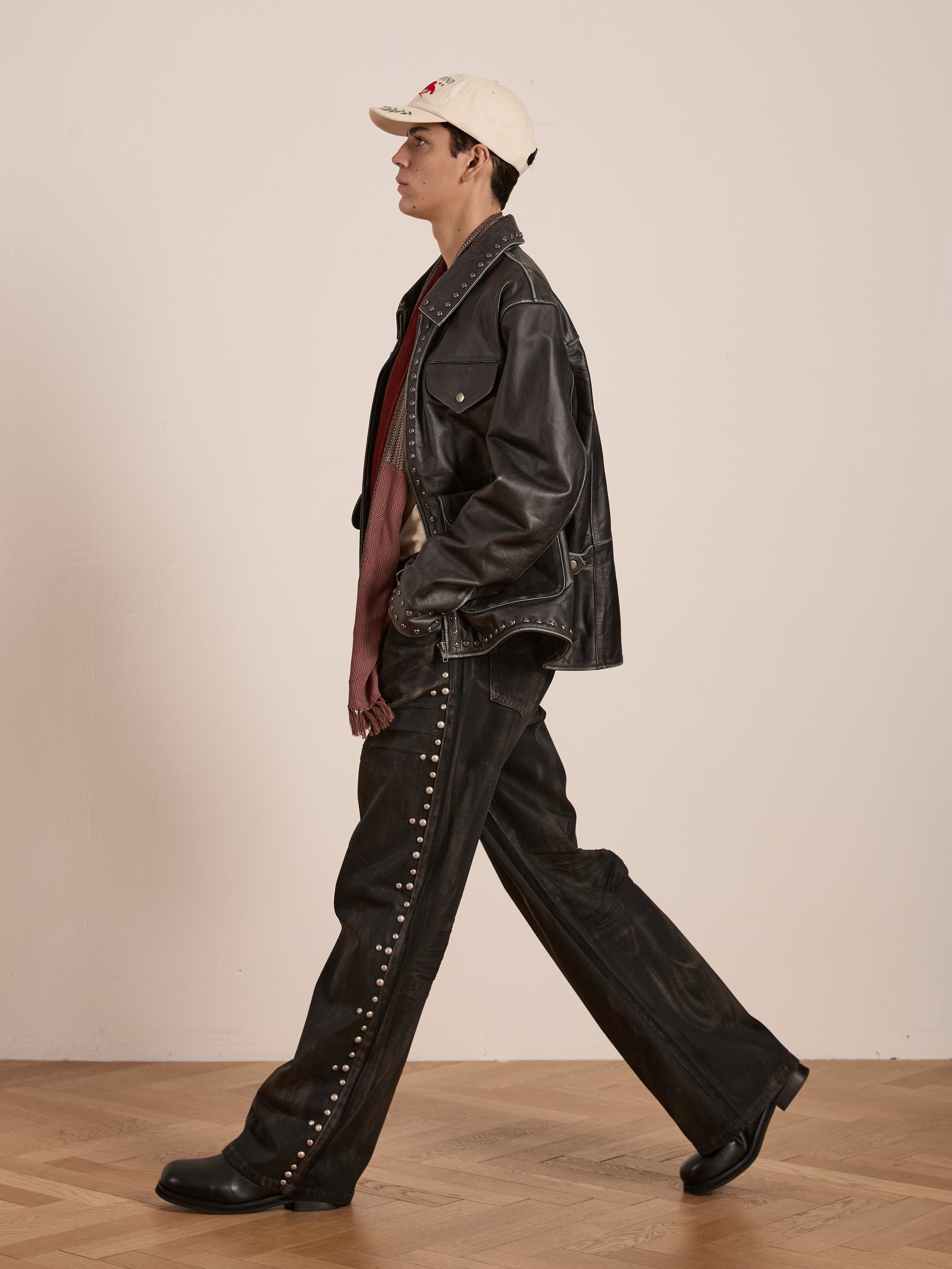 Clad in Found's Studded Leather Pocket Jacket and matching pants, the individual strides sideways. A beige cap completes the look as they move across the wooden indoor floor.