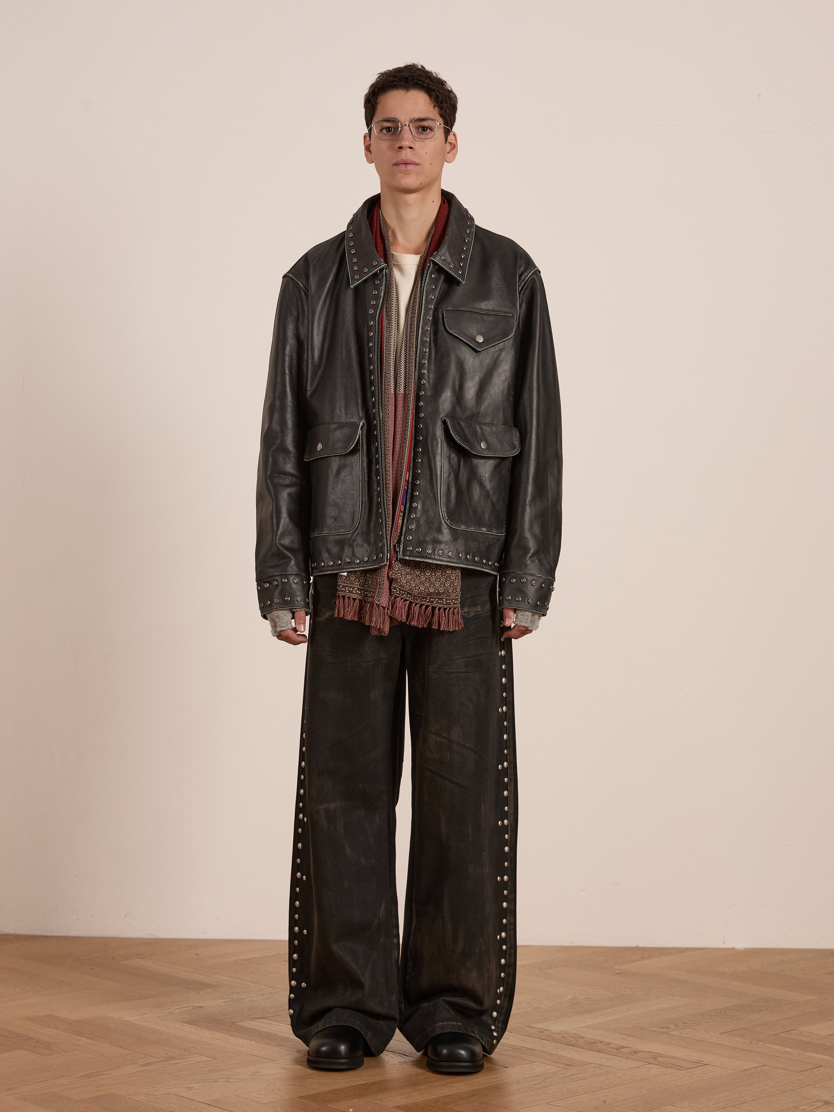 A model poses in Found's vintage Studded Leather Pocket Jacket and a red scarf with wide-legged studded pants. The setting includes a plain, light-colored wall and wooden floor.