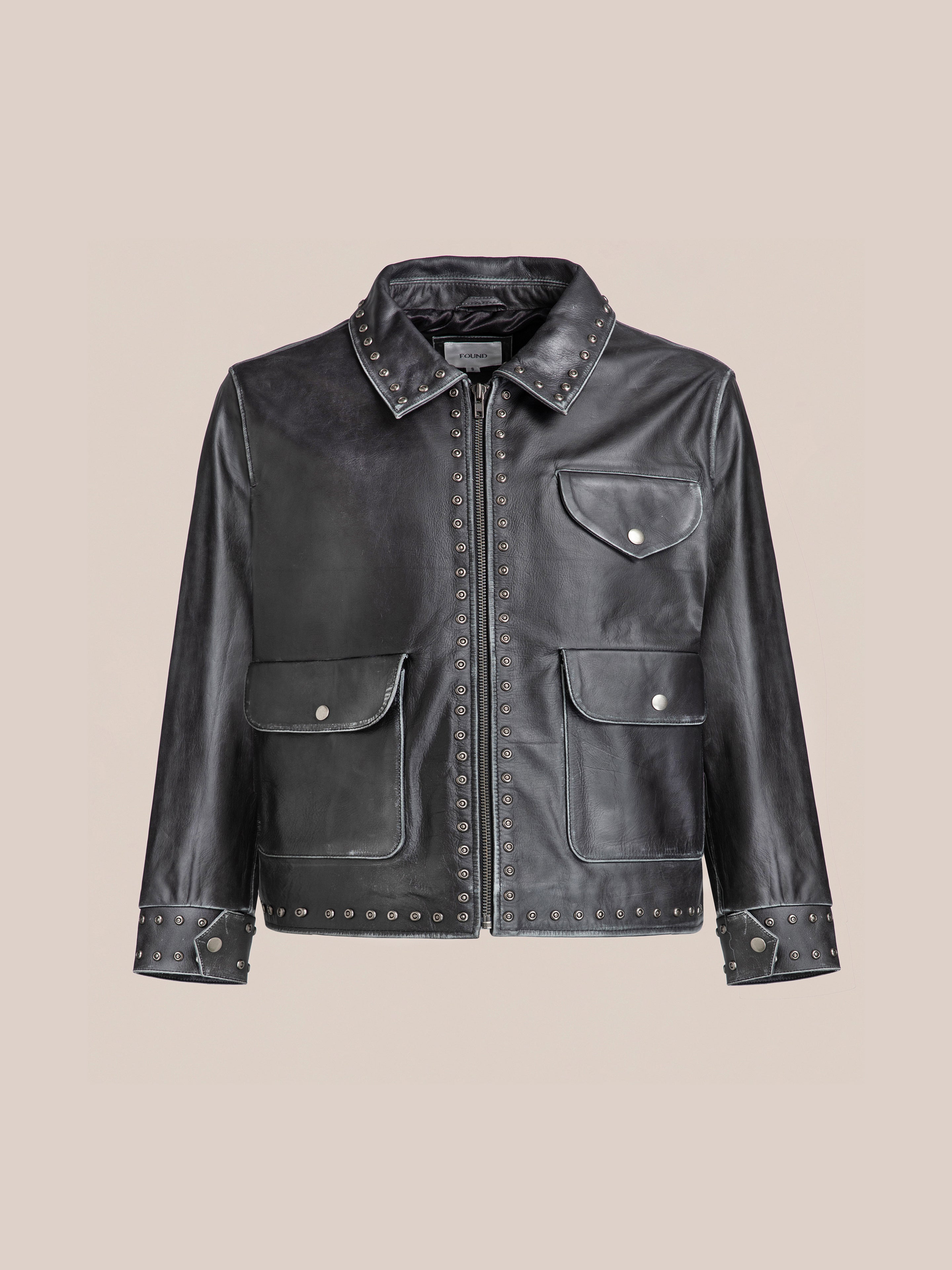 The Studded Leather Pocket Jacket by Found is a vintage black leather piece with silver studs, a front zipper, two large pockets, a chest pocket, and studded cuffs—all set on a beige background for a timeless yet edgy unisex statement.