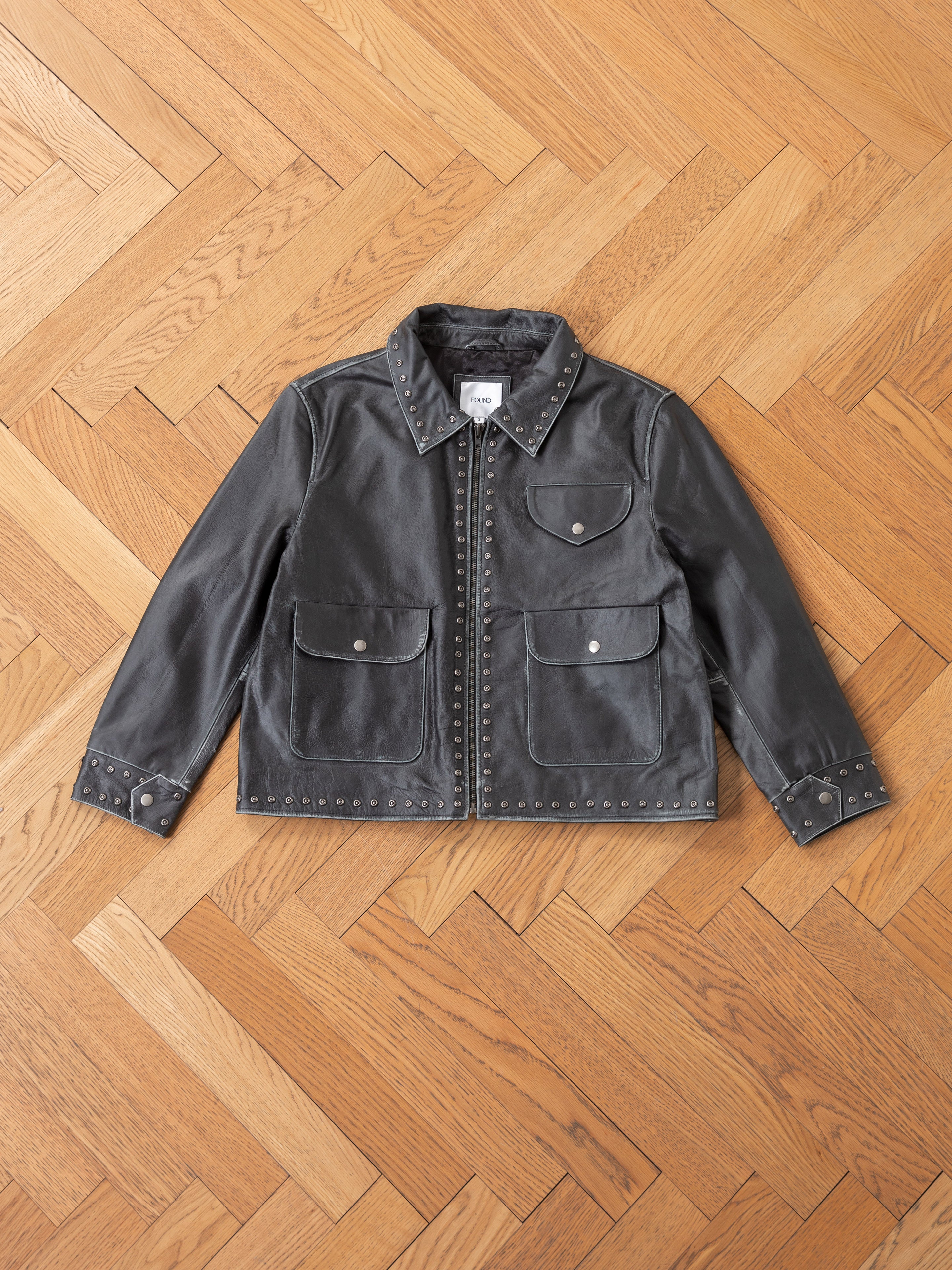 The Studded Leather Pocket Jacket by Found, a vintage-style black leather piece adorned with silver studs, elegantly rests on a wooden herringbone floor, exuding timeless style.