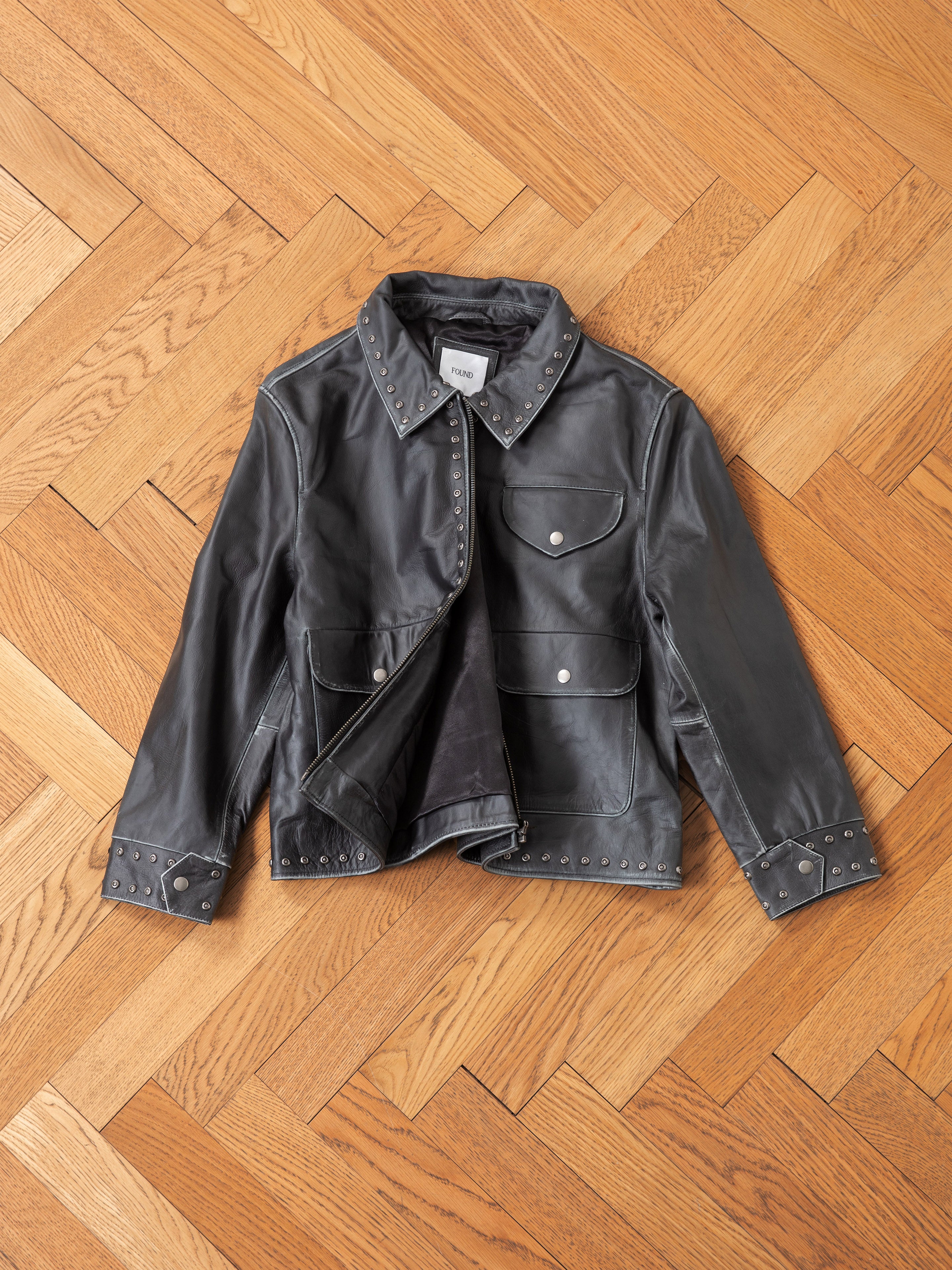 The Studded Leather Pocket Jacket by Found, a vintage black leather piece with silver studs on the collar, pockets, cuffs, and hem, is laid on a wooden herringbone-patterned floor.