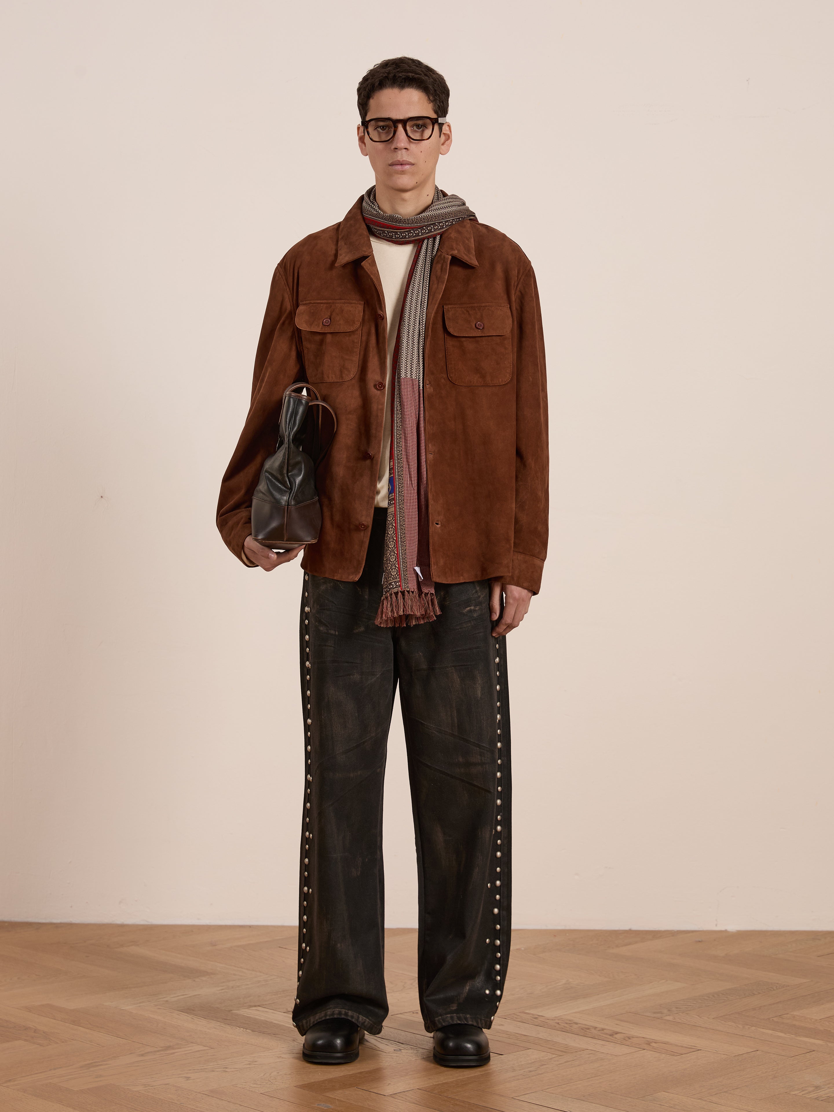 A person wears the FOUND Suede Camp Shirt, echoing a classic camp style, paired with wide dark studded pants, a scarf, and glasses. They hold a dark bag while standing on a wooden floor.