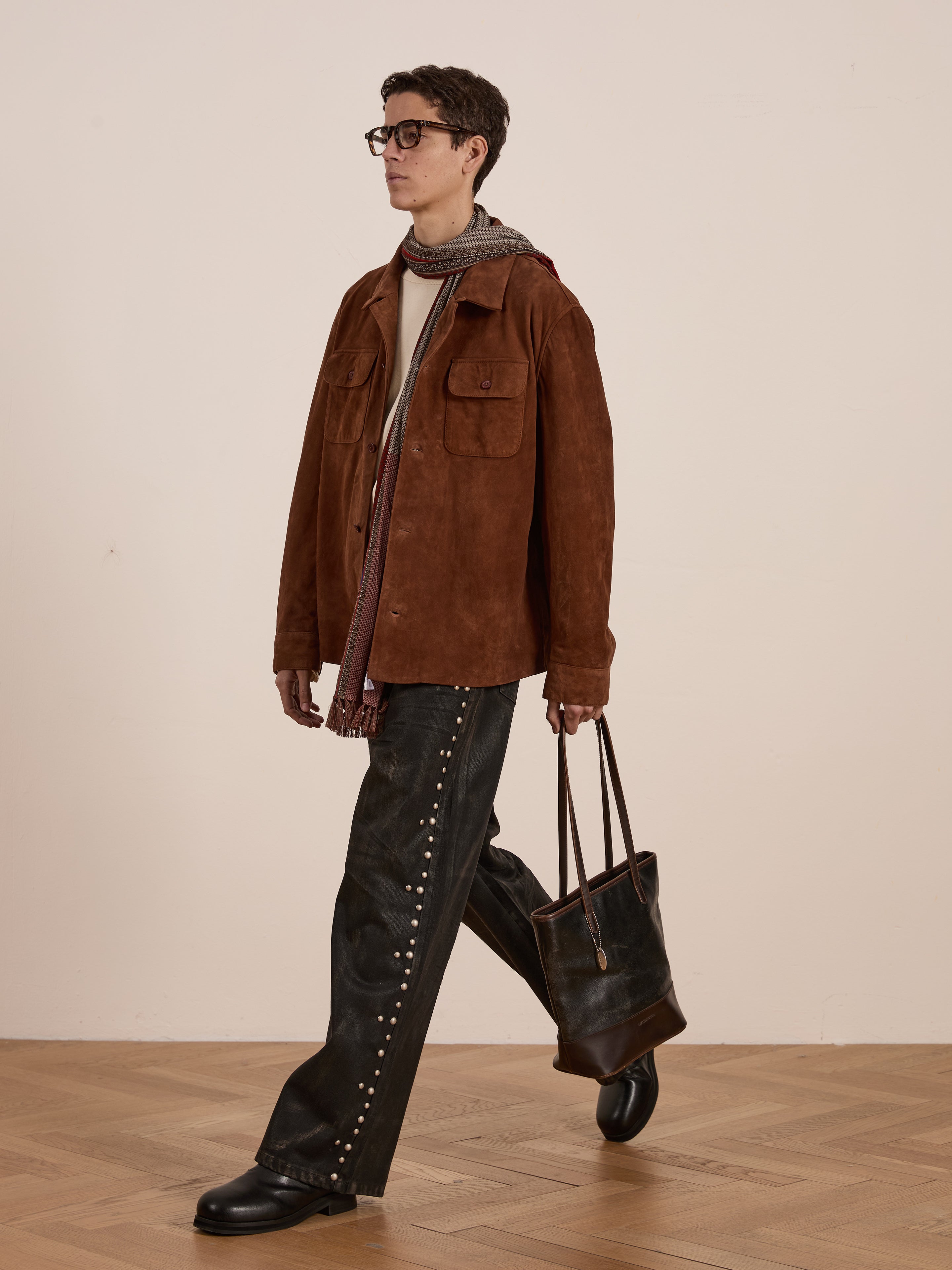 Dressed in the FOUND Suede Camp Shirt and a gray scarf, paired with studded baggy jeans, they stroll on a wooden floor with a dark tote bag, showcasing effortless style.