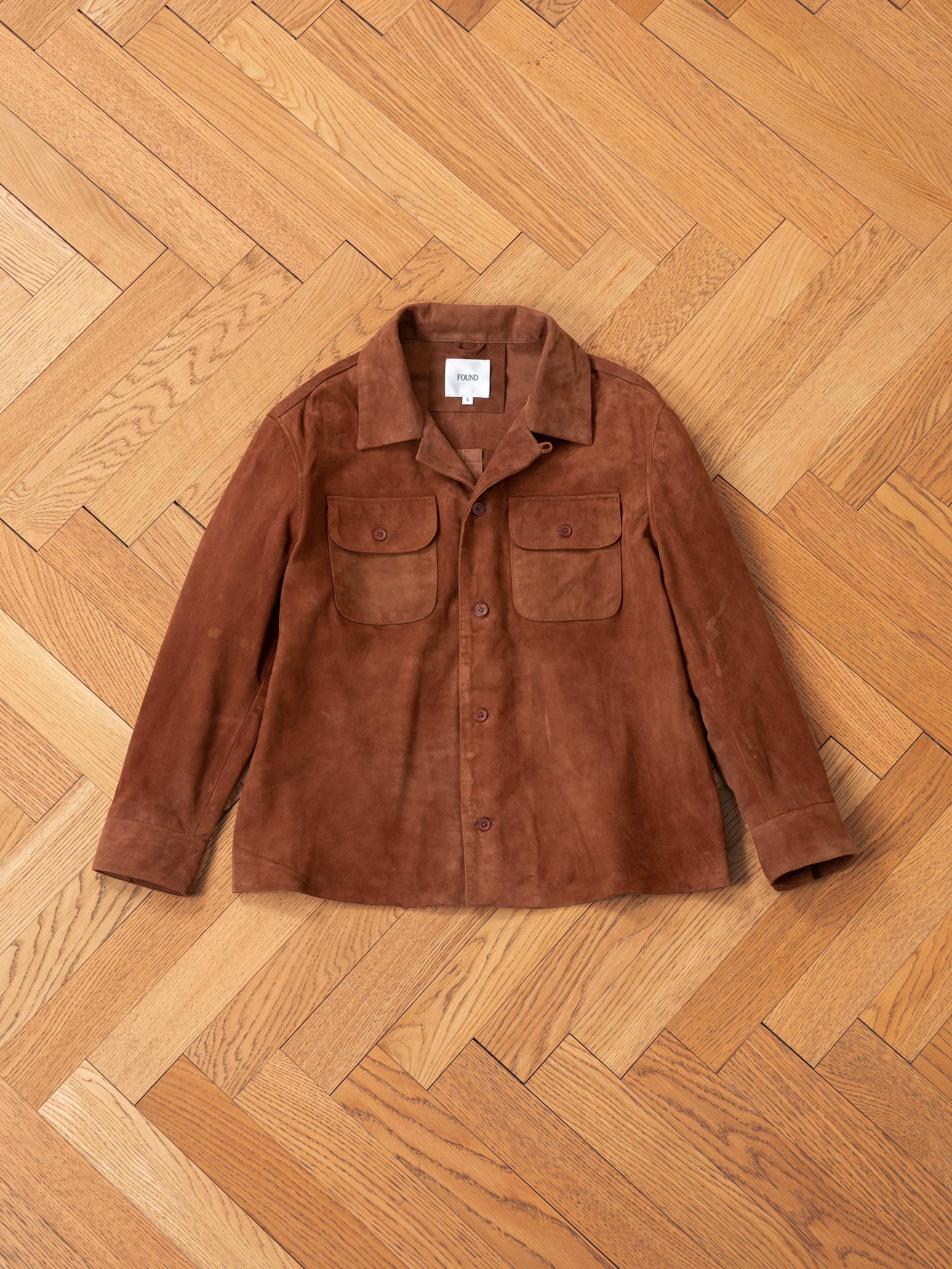 The FOUND Suede Camp Shirt in brown features a relaxed fit, button-up design, collar, and two chest pockets. Made from genuine goat suede leather, it elegantly complements the wooden herringbone floor.