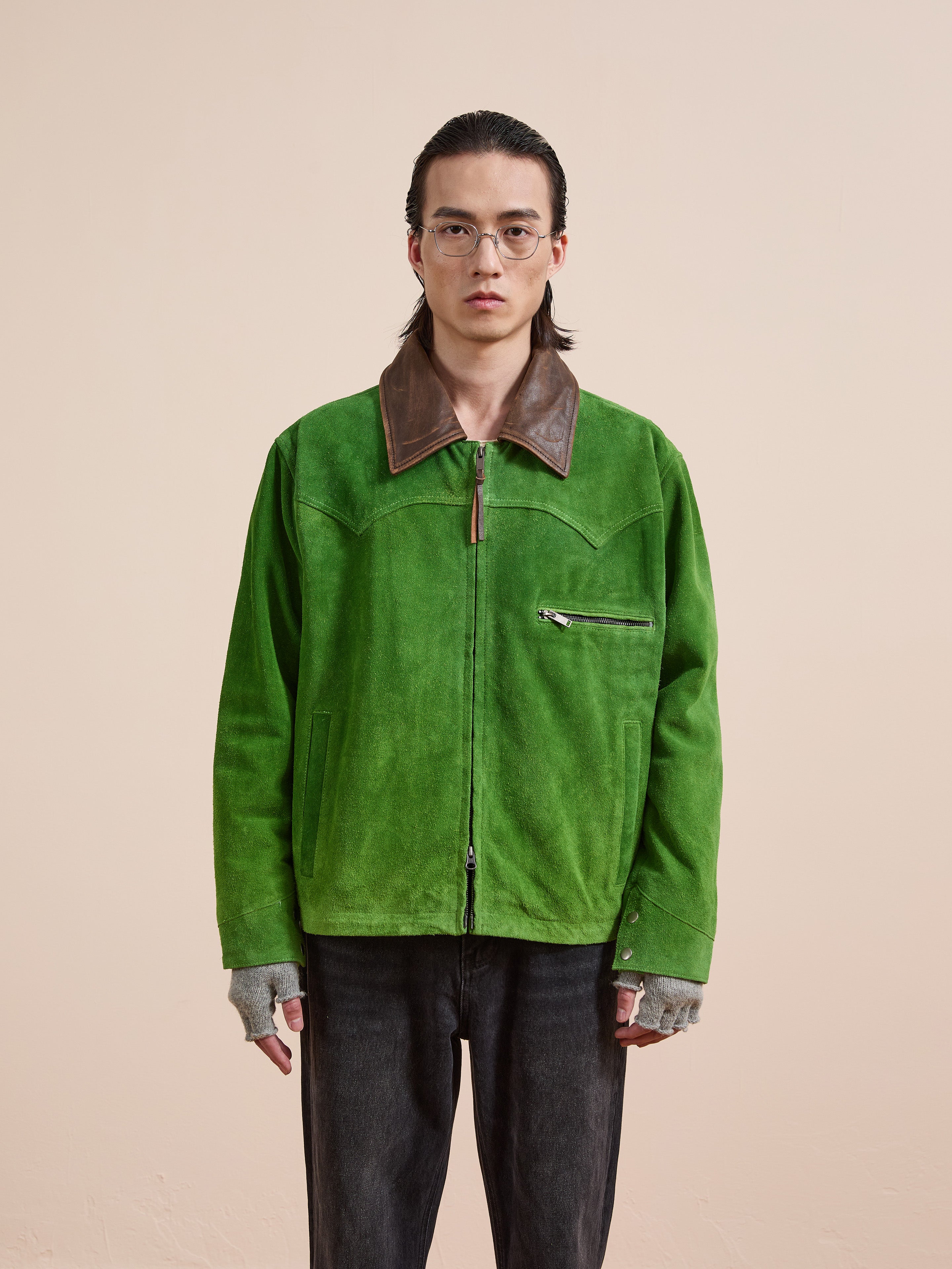 A person with long hair, glasses, and fingerless gloves wears the FOUND Suede Western Barn Jacket—a green genuine suede jacket with a brown collar and western yoke stitching—against a plain beige background.