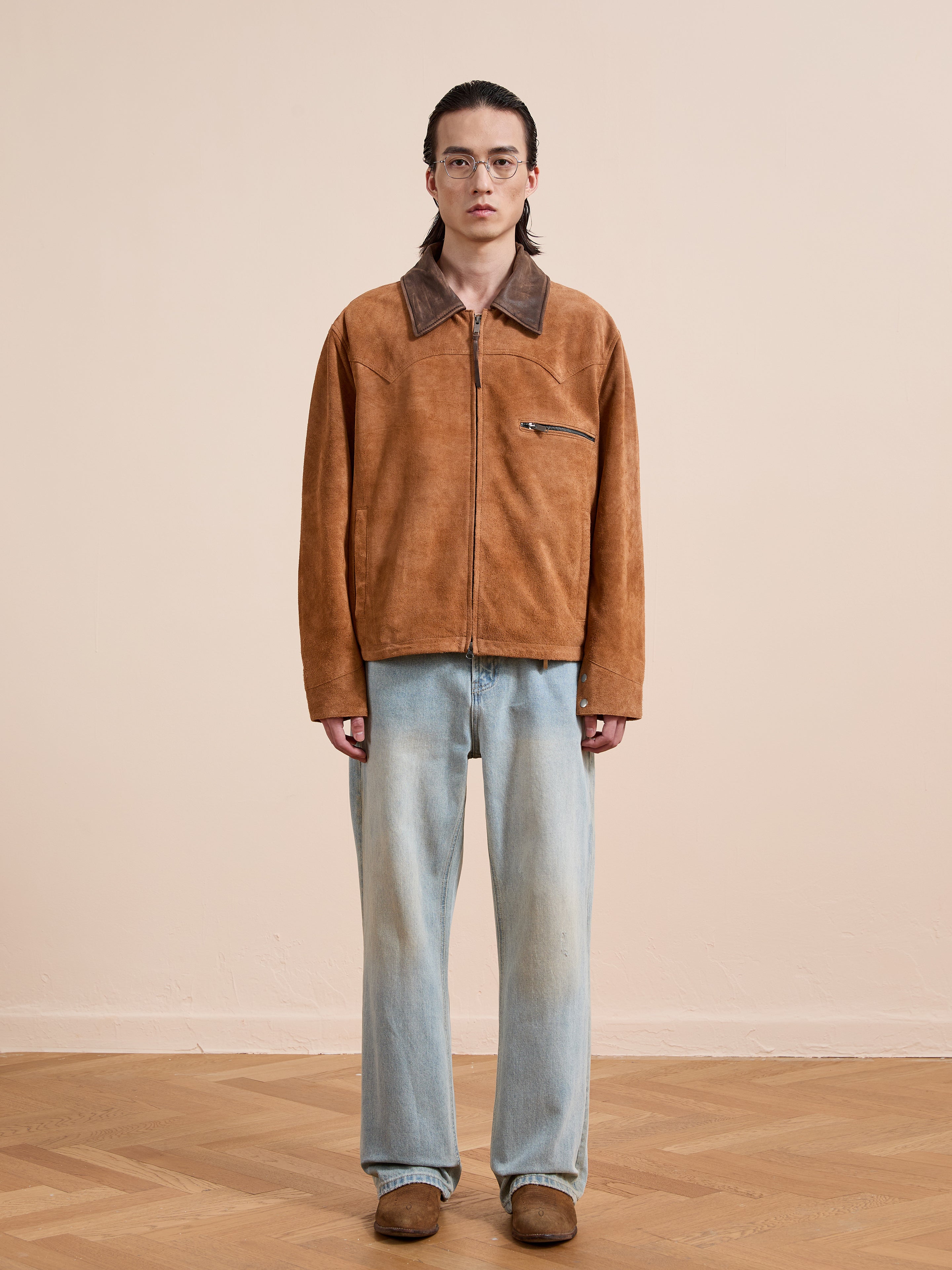 A person stands indoors against a beige wall and wooden floor, wearing FOUND's Suede Western Barn Jacket with western yoke stitching, paired with light blue jeans, brown shoes, and glasses.