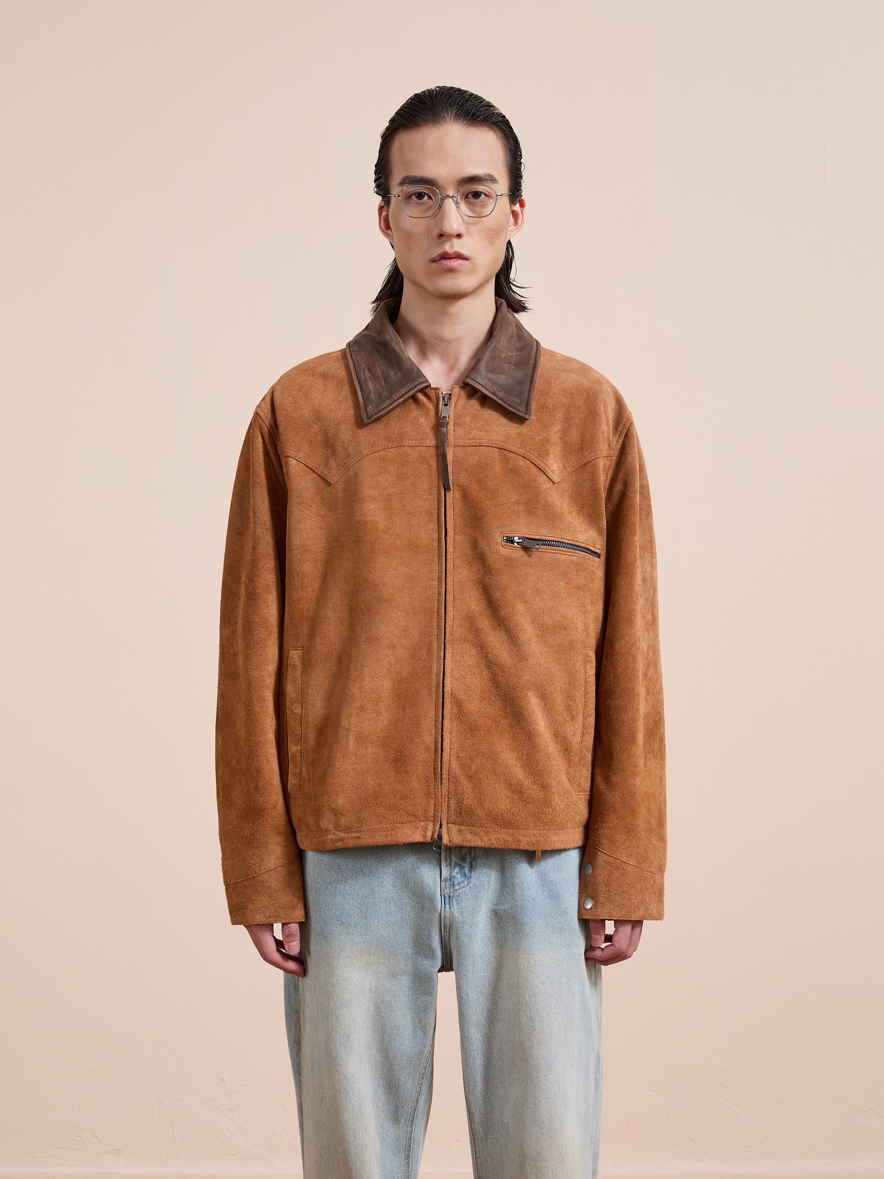 A person with long dark hair and glasses wears the FOUND Suede Western Barn Jacket and light blue jeans, standing against a plain beige background.