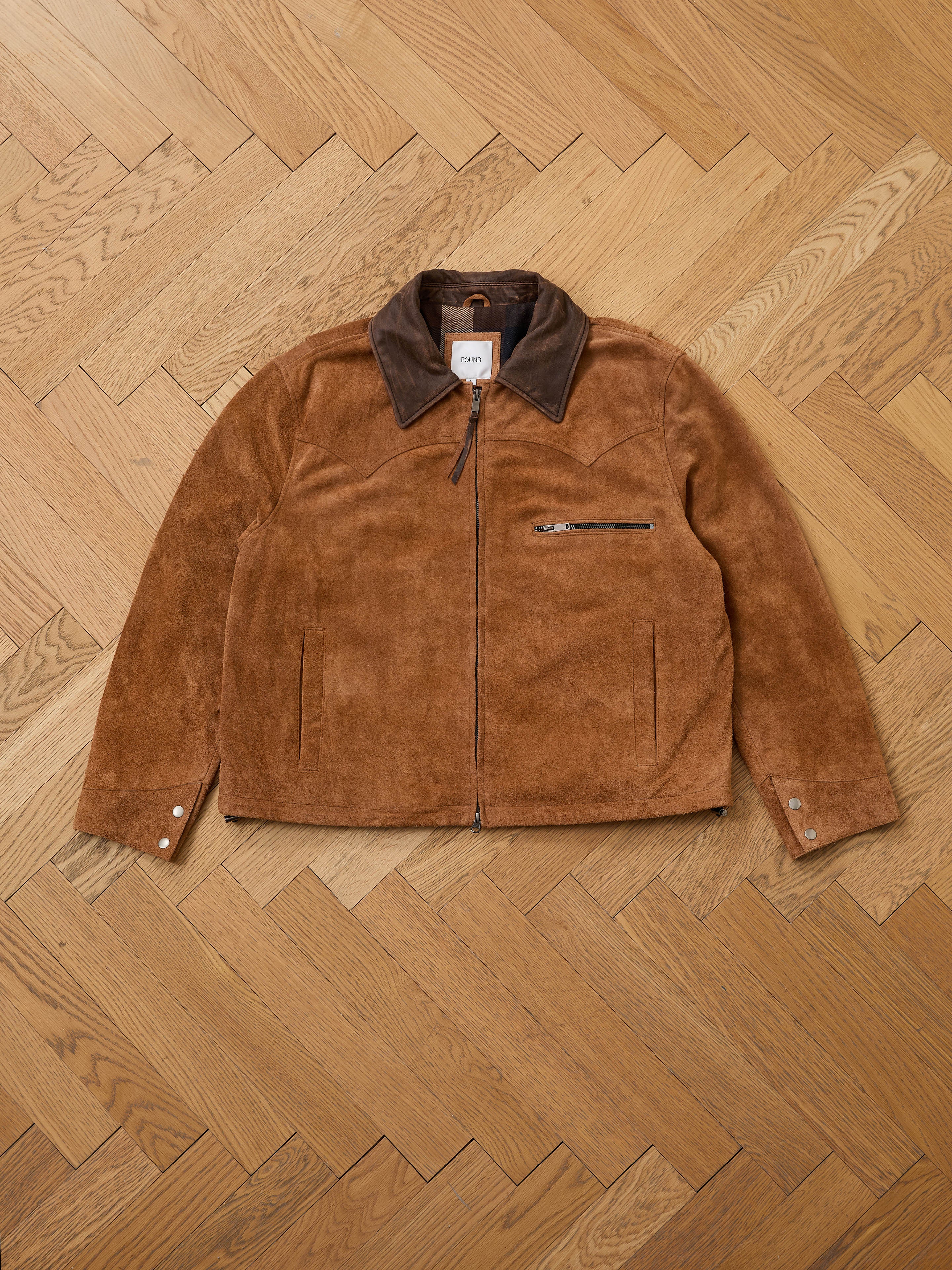 The FOUND Suede Western Barn Jacket features a brown suede finish, dark collar, western yoke stitching, zip front, chest zip pocket, and two side pockets, displayed flat on a herringbone-patterned wooden floor.