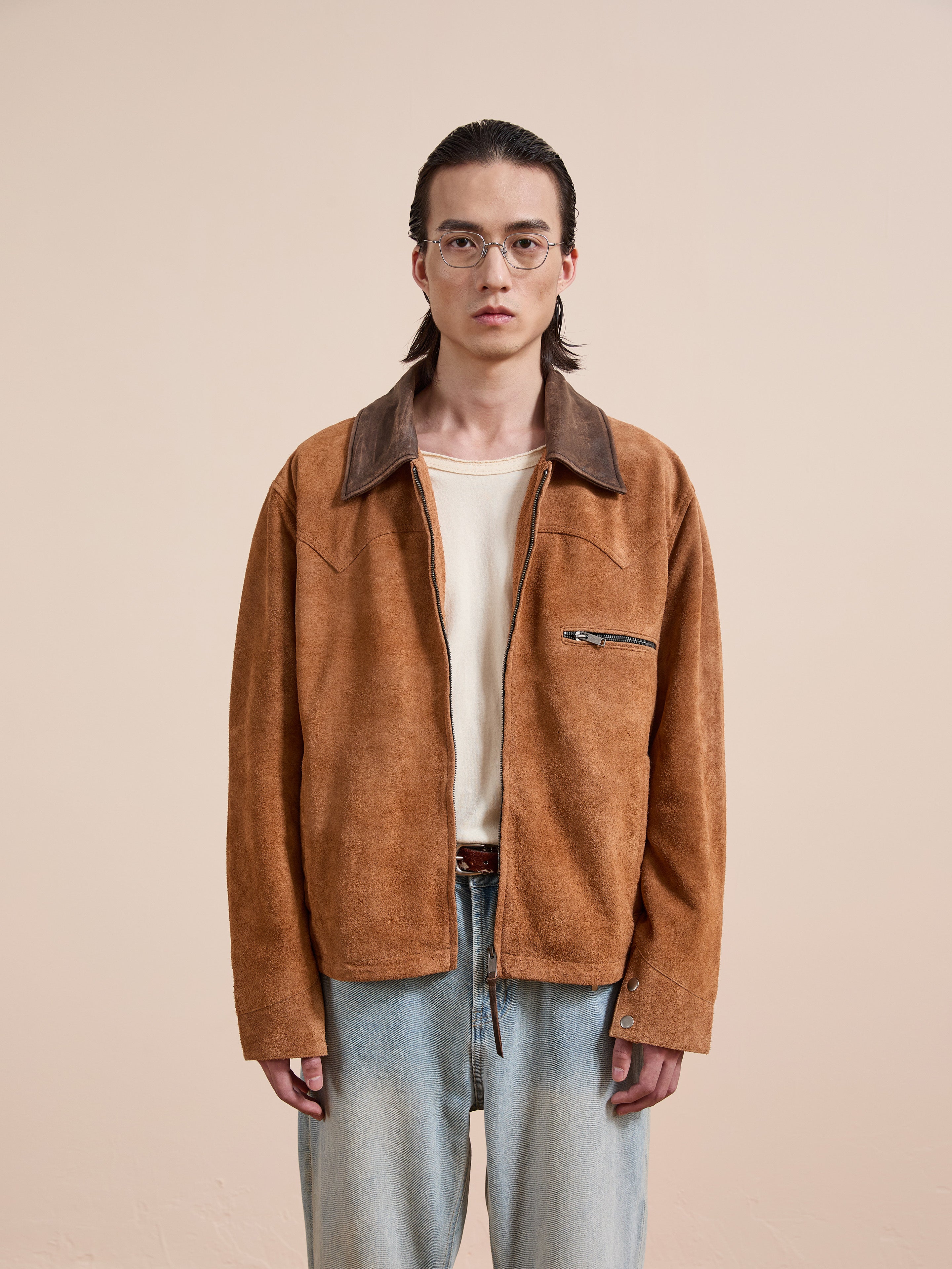 A person with long dark hair and glasses wears the FOUND Suede Western Barn Jacket over a cream shirt and light blue jeans, standing against a plain beige background.