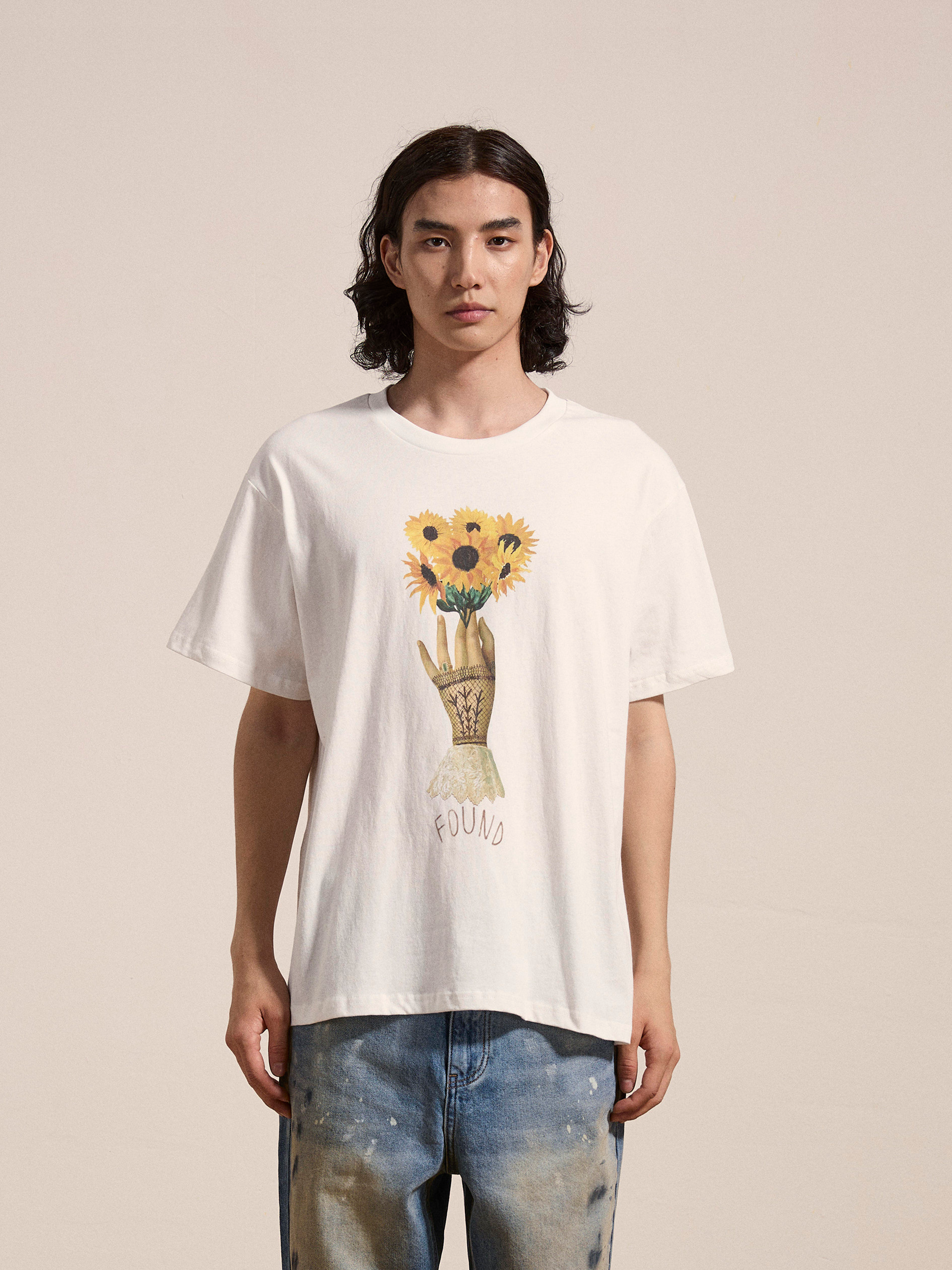 A person with long hair is wearing a white Sunflower Tee from FOUND, featuring a floral graphic of a hand holding sunflowers with the word "FOUND" below it. They are also wearing blue jeans.