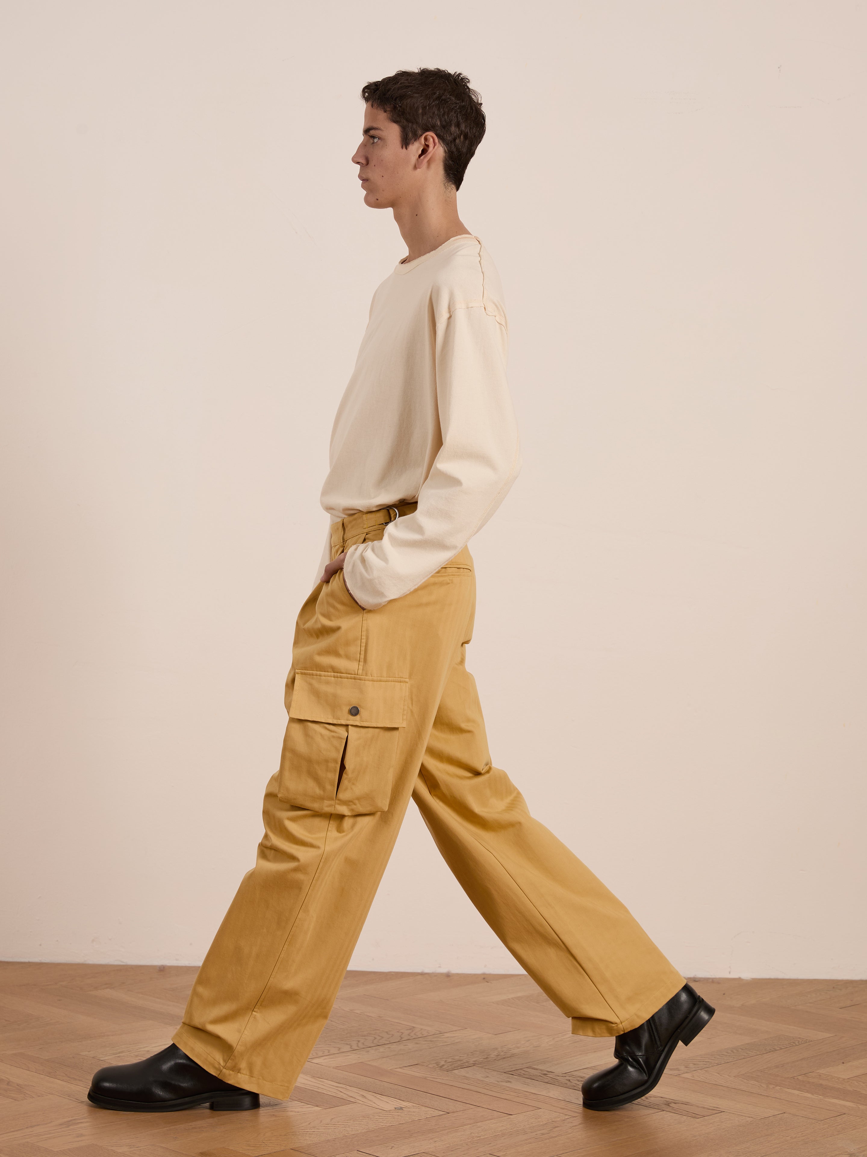 A person wearing a cream long-sleeve shirt and FOUND's Sunlight Twill Cargo Pants in a relaxed fit walks in profile on a wooden floor.