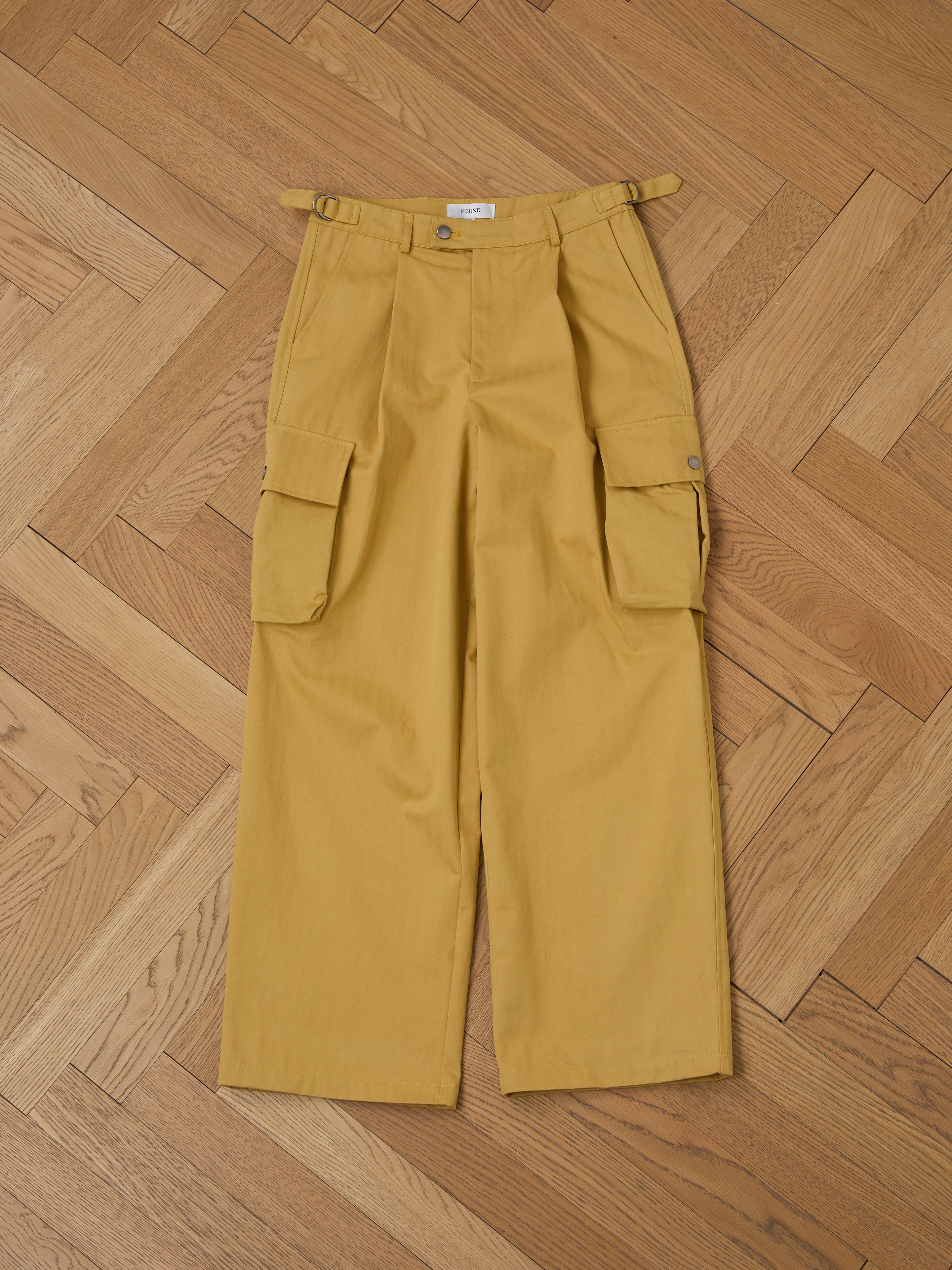 The Sunlight Twill Cargo Pants by FOUND, featuring a unisex design with wide-leg, relaxed fit, side pockets, and adjustable waist tabs, are shown laid flat on a wooden herringbone floor.