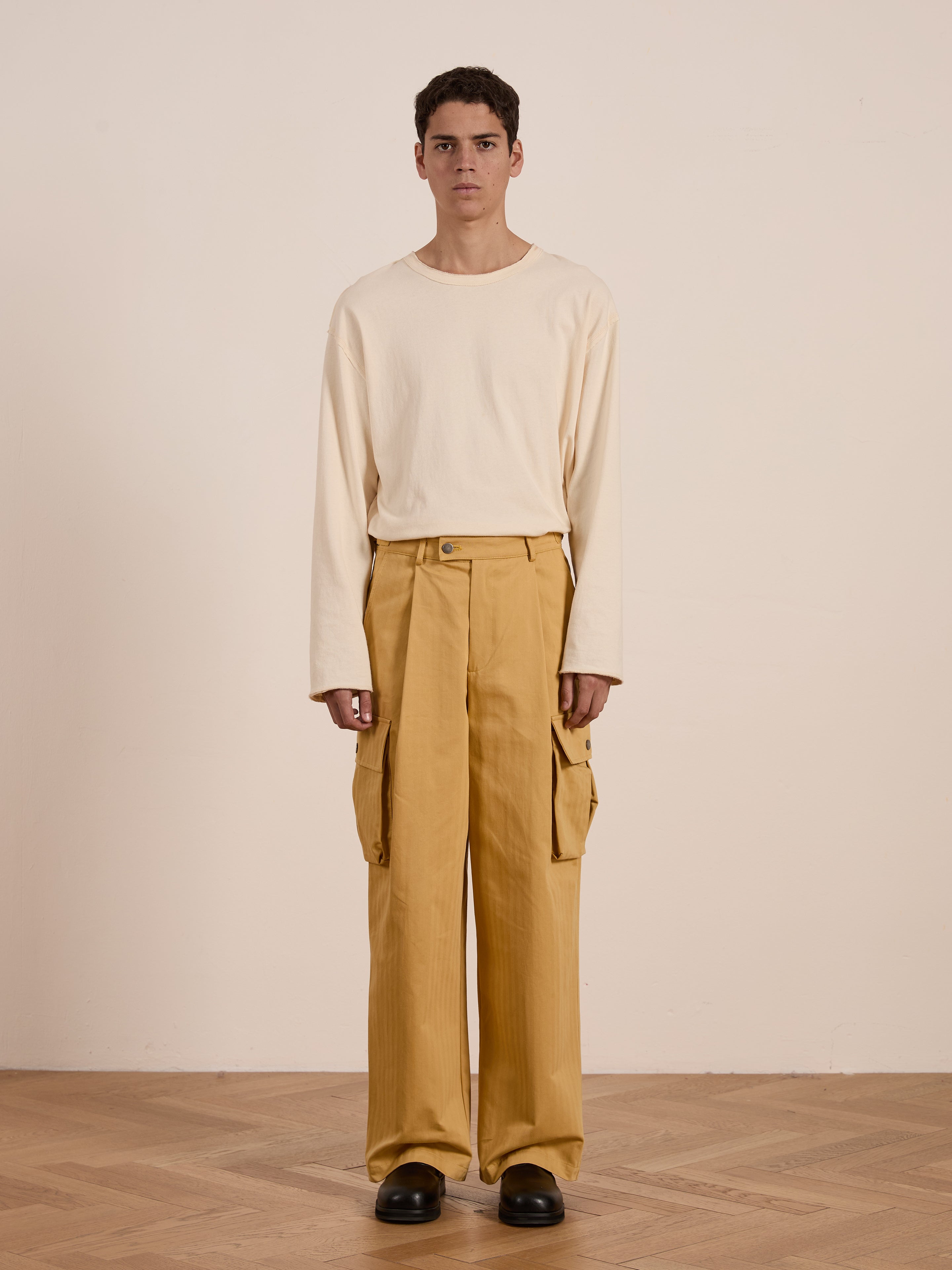 Indoors on a wooden floor against a plain background, someone is casually dressed in a cream long-sleeve shirt and FOUND's Sunlight Twill Cargo Pants.