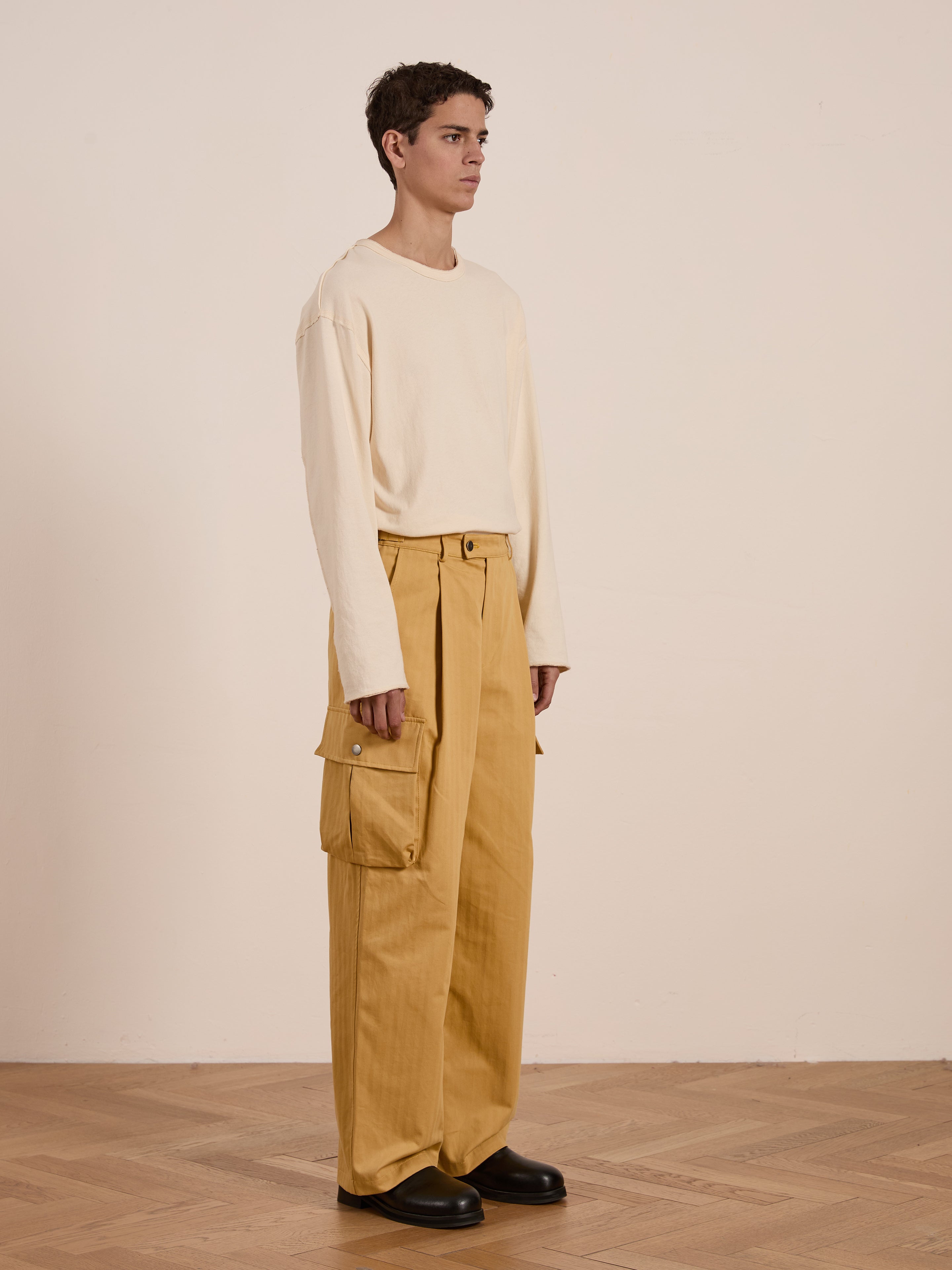 In a minimalist setting, an individual dons a cream long-sleeve shirt with relaxed fit, mustard-colored Sunlight Twill Cargo Pants from FOUND, showcasing large pockets. Black shoes finish this unisex ensemble.