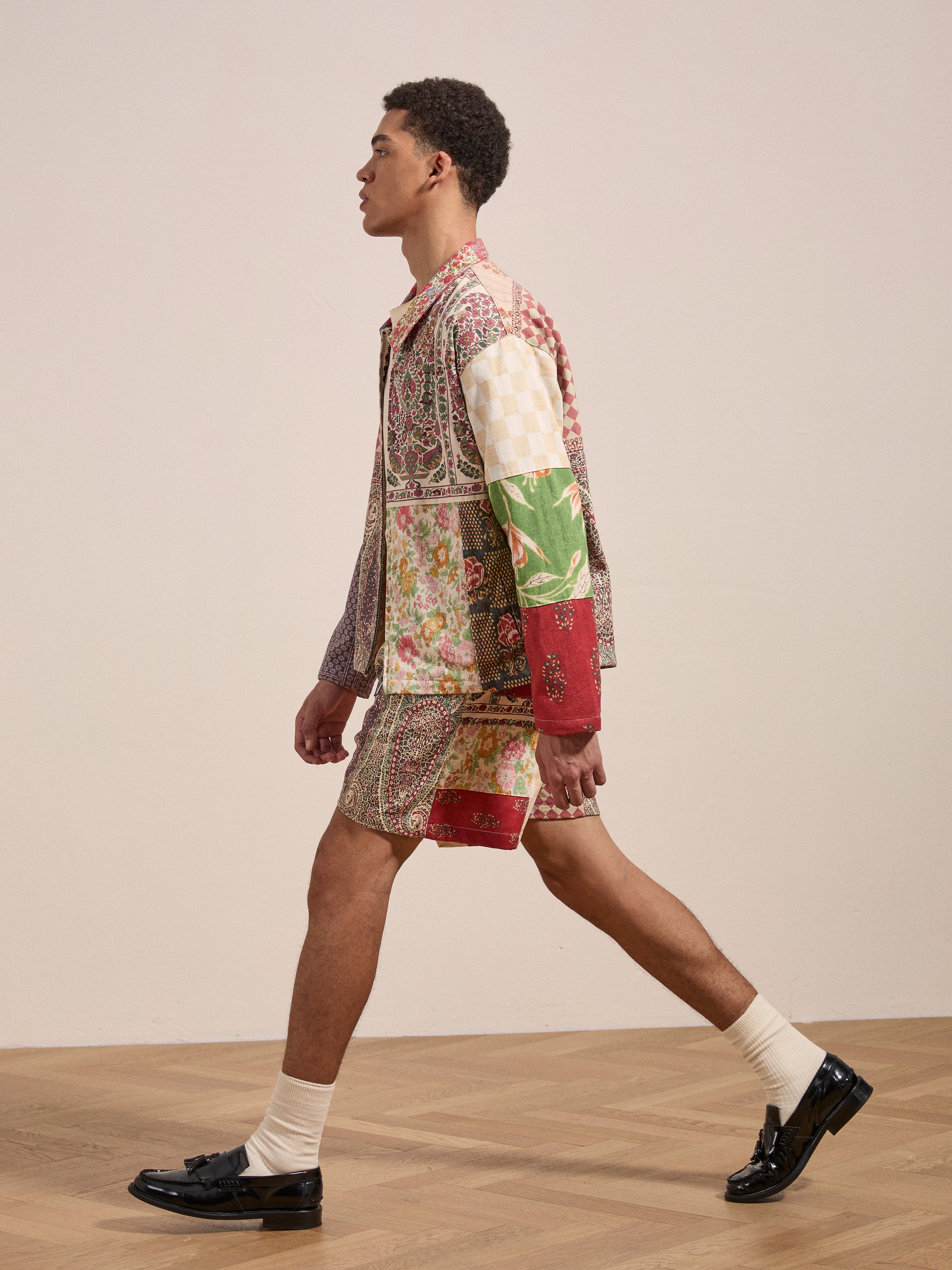 A man wearing the Tapestry Twill Shorts by FOUND, featuring various patterns in a patchwork design, walks in profile on a wooden floor, paired with white socks and black loafers.