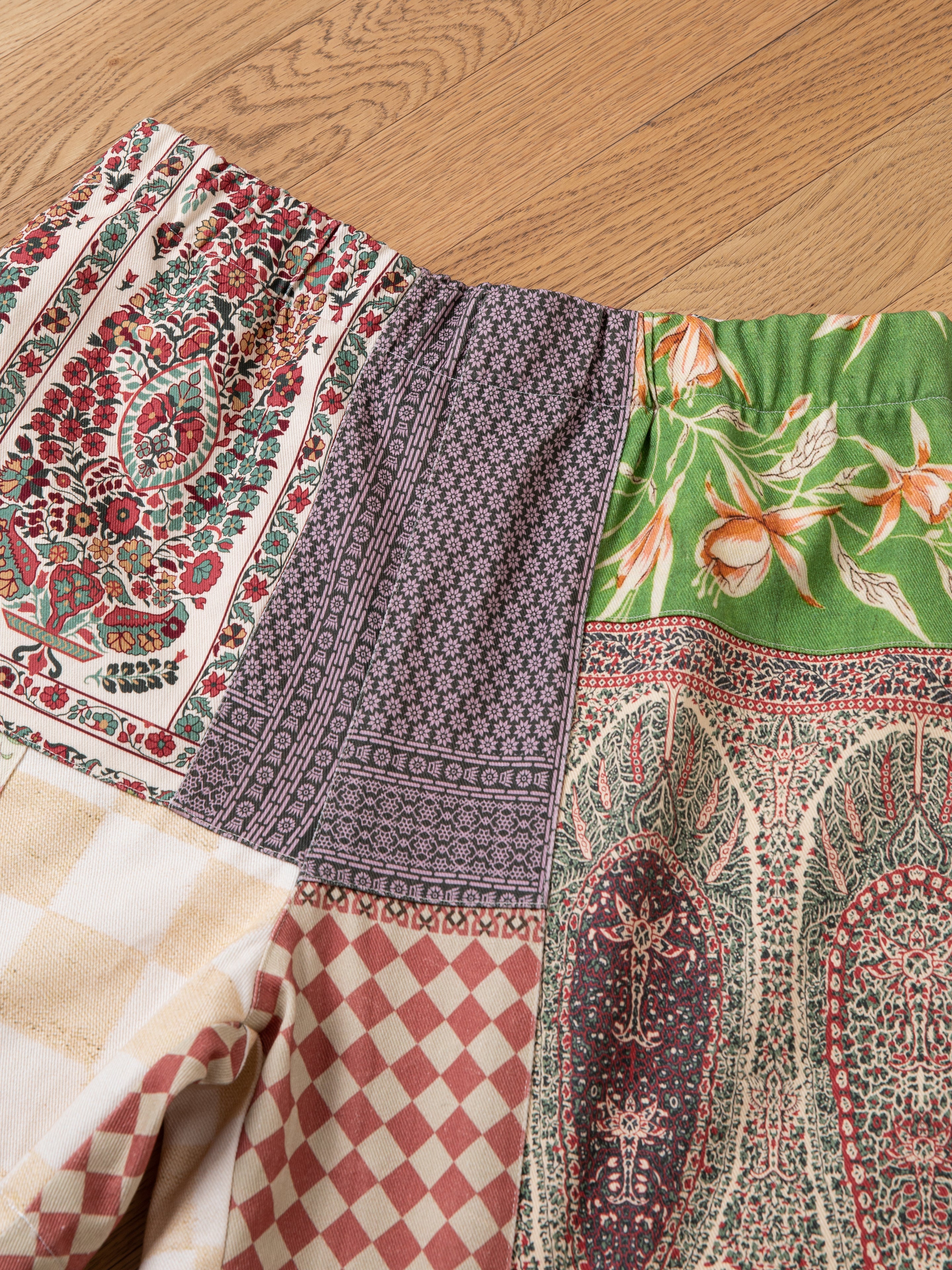 Close-up of the FOUND Tapestry Twill Shorts, showcasing a patchwork design with various colorful patterns, including florals, geometrics, and paisleys, laid out on a textured wooden floor.