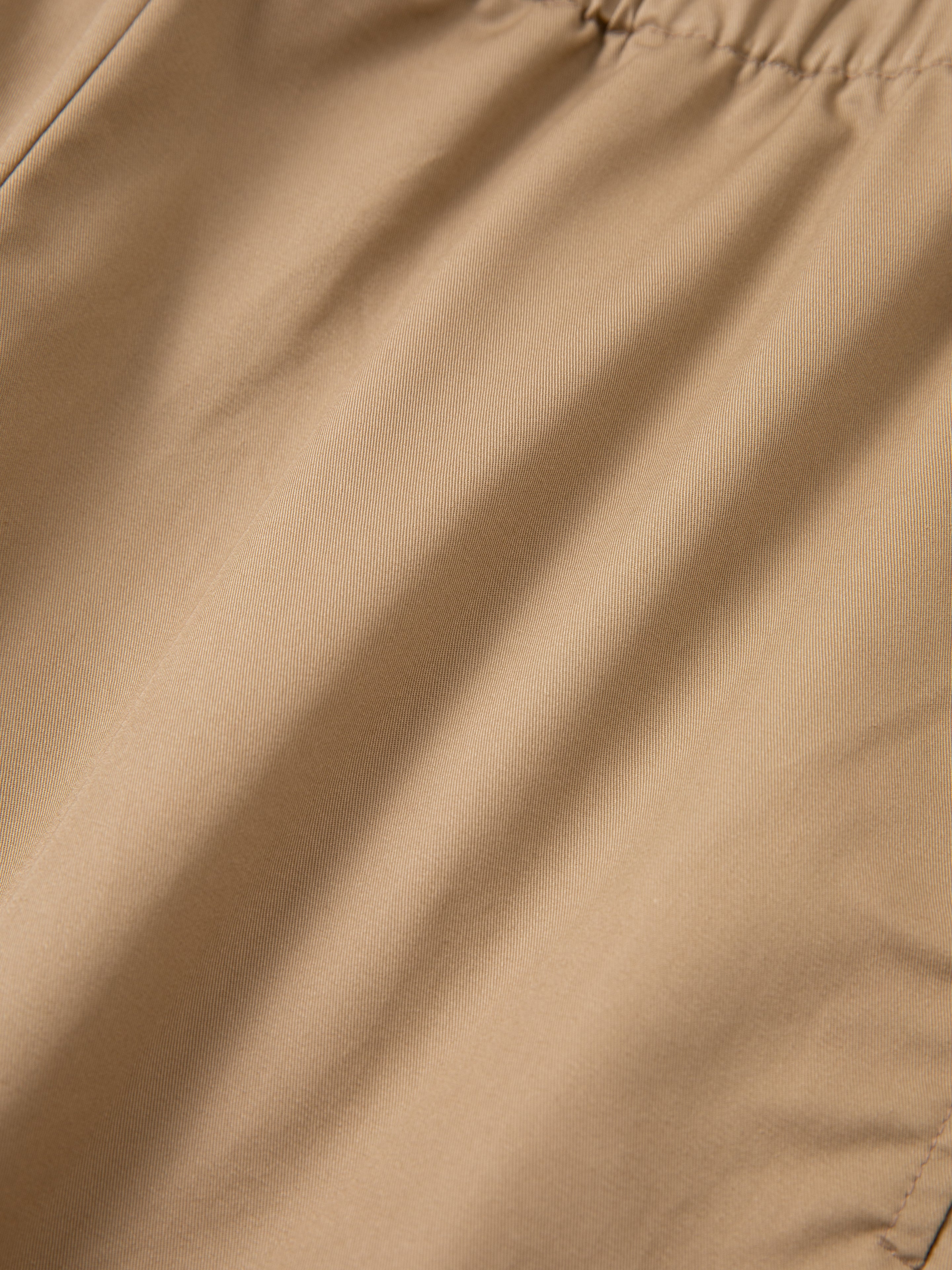 Here is a close-up of light brown, lightweight cotton fabric with a smooth texture and slightly visible weave pattern from the Long Twill Cargo Shorts by FOUND. The fabric exhibits a faint sheen and appears to be slightly gathered or wrinkled, making it ideal for twill cargo drawstring shorts equipped with dual cargo pockets.