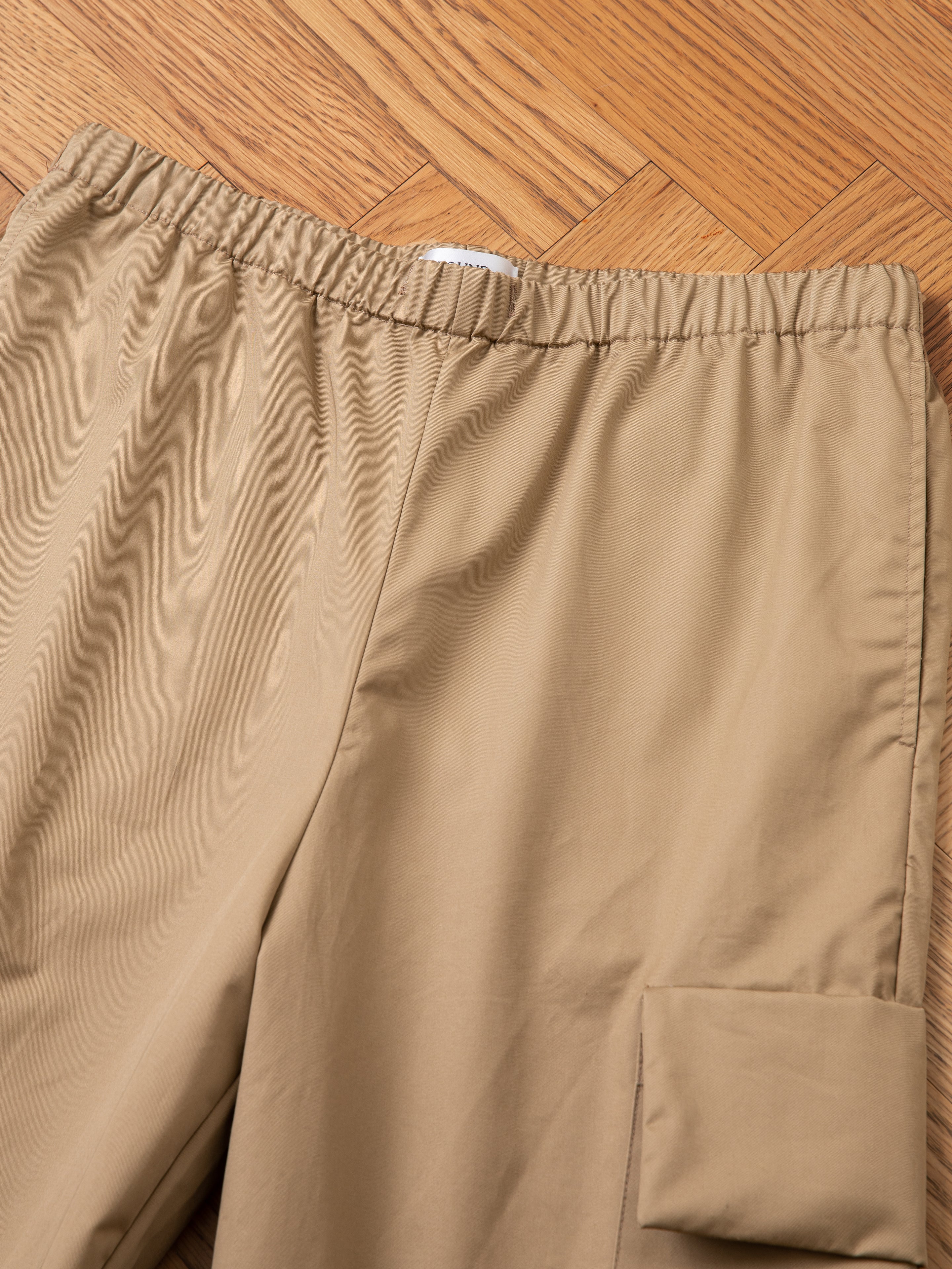 Close-up of beige Long Twill Cargo Shorts by FOUND, featuring an elastic waistband and dual cargo pockets, laid on a wooden floor.