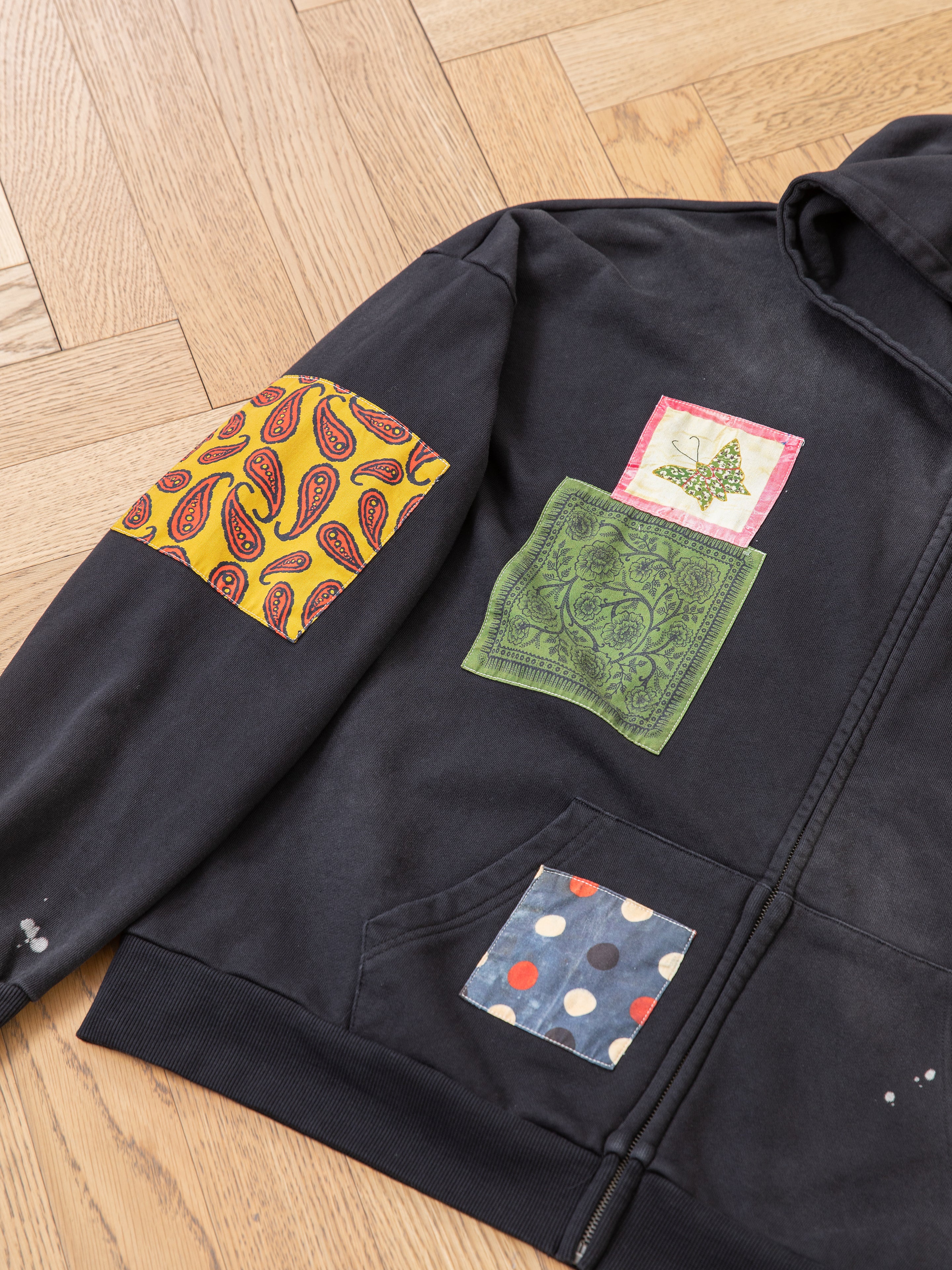 The Tapestry Patchwork Zip Up Hoodie by FOUND showcases a vibrant design with colorful patches: a yellow paisley adorns the sleeve, a unique green paisley with an animal motif highlights the chest, and blue fabric with white dots decorates the pocket. Its vintage-inspired tapestry aesthetic is beautifully displayed on a light wood floor.