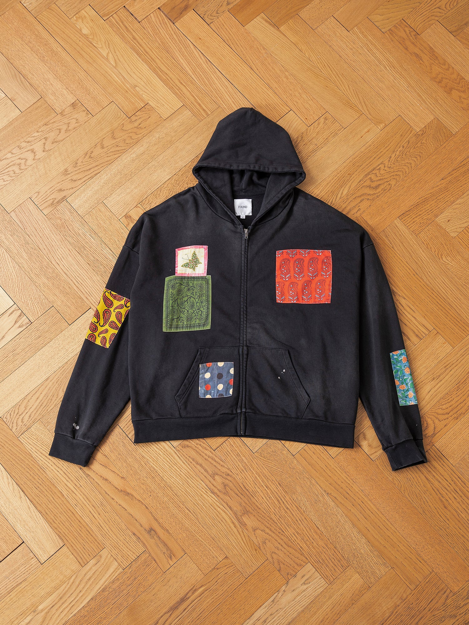 The Tapestry Patchwork Zip Up Hoodie by FOUND features a vintage-inspired oversized fit, embellished with colorful patchwork designs and beautifully displayed on a wooden herringbone-patterned floor.