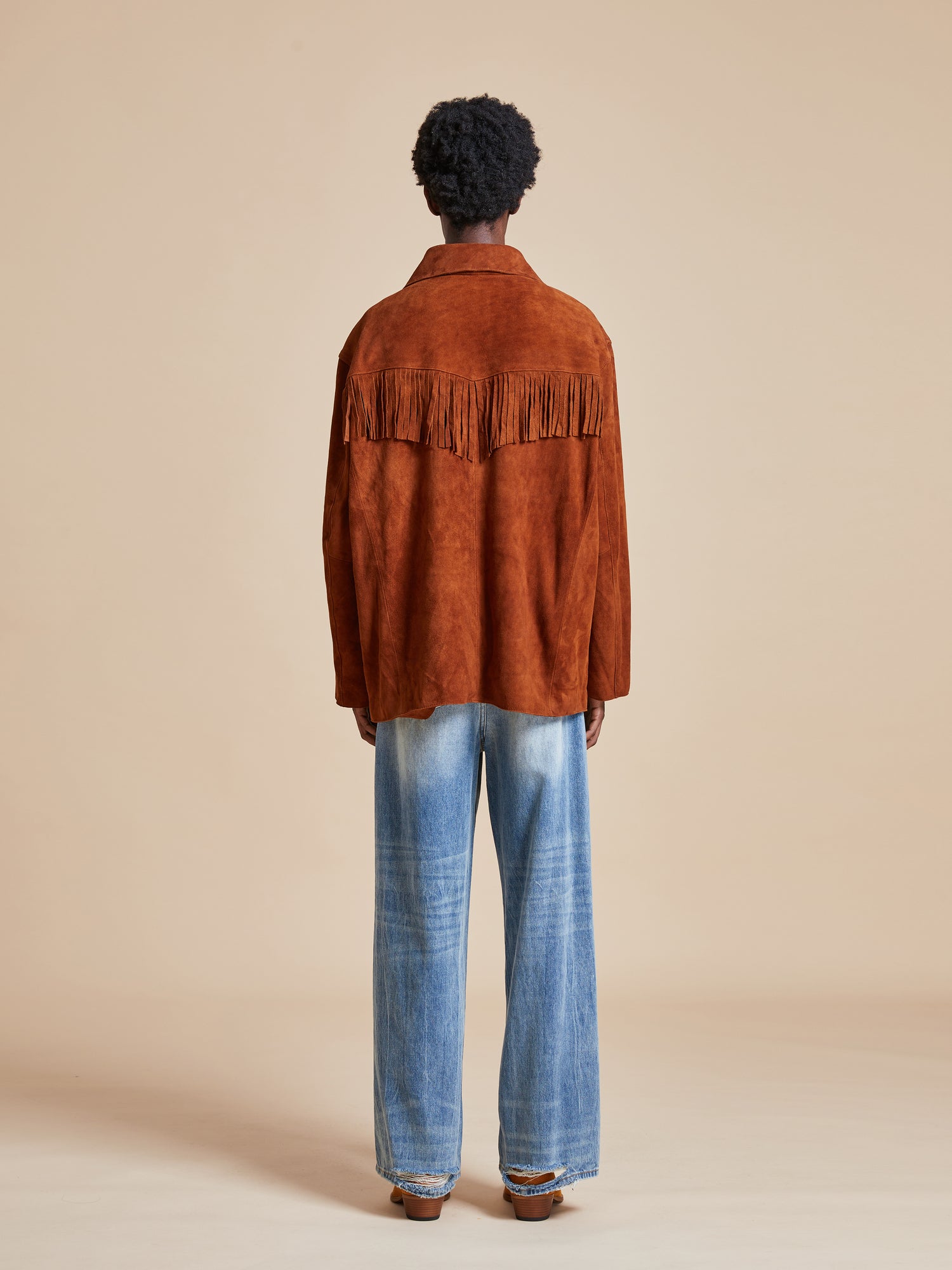 The back view of a man wearing a Found Tobacco Fringe Suede Buckle Jacket and jeans.