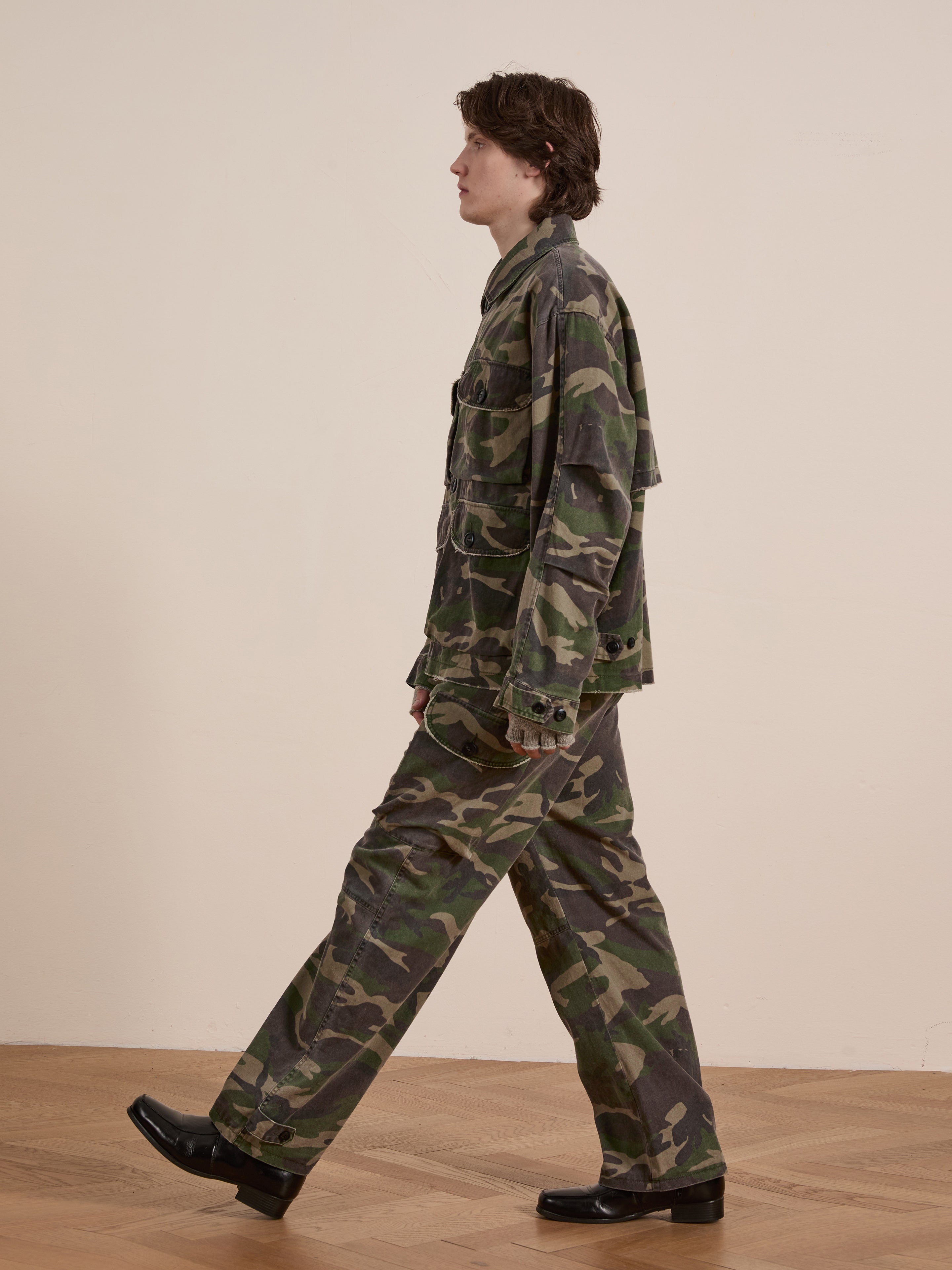 Wearing the FOUND Camouflage Twill Utility Cargo Jacket, a person strolls across the wooden floor, perfectly complementing their vintage look as they face left.