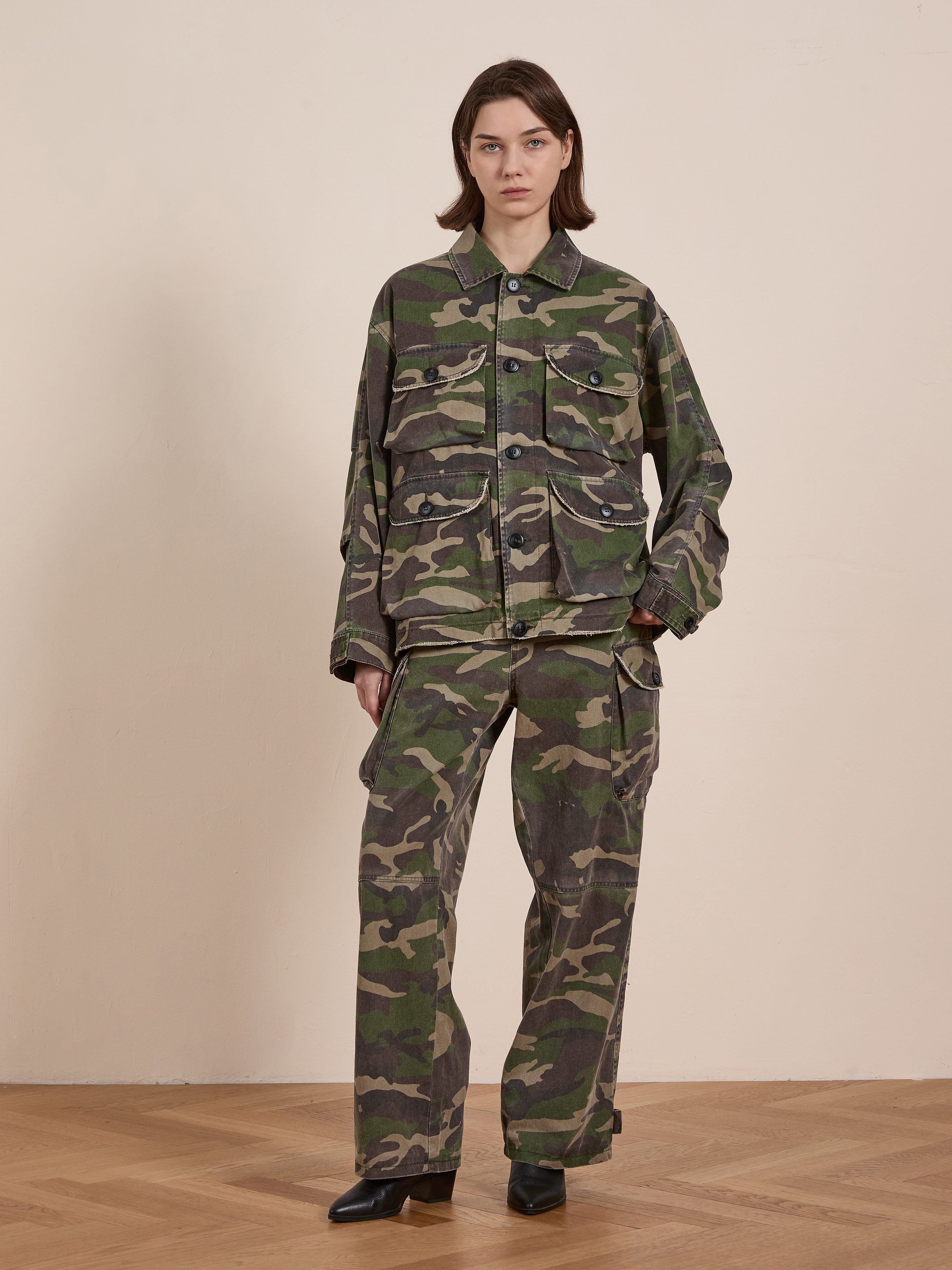 A person wears the FOUND Camouflage Twill Utility Cargo Jacket with matching pants, standing on a wooden floor against a plain wall, effortlessly capturing a vintage look.