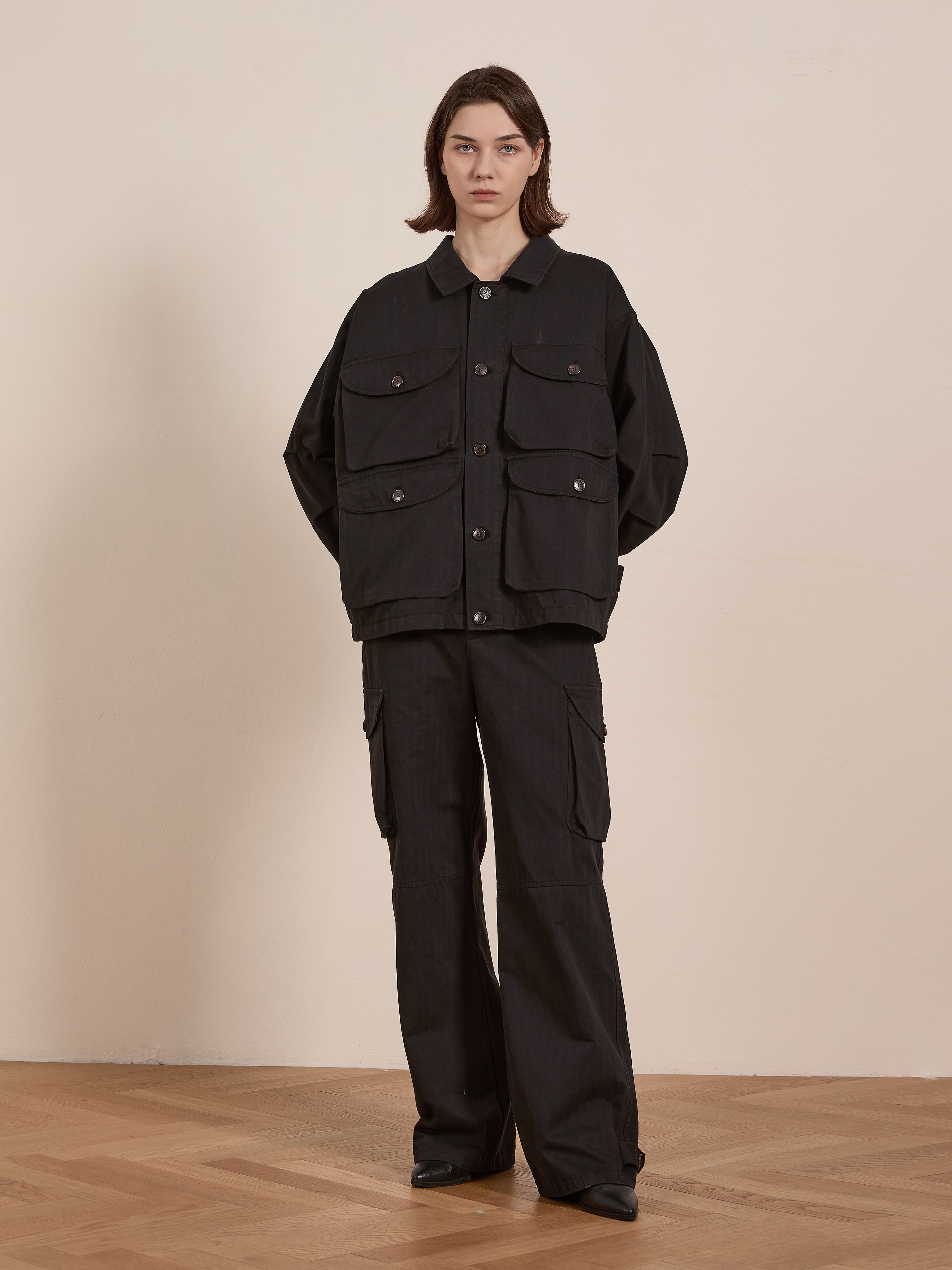 Indoors, a person sports a loose-fitting black Utility Herringbone Cargo Jacket by FOUND with matching wide-leg pants, capturing a vintage look. The wooden floor and beige background wall amplify the timeless style of the outfit.