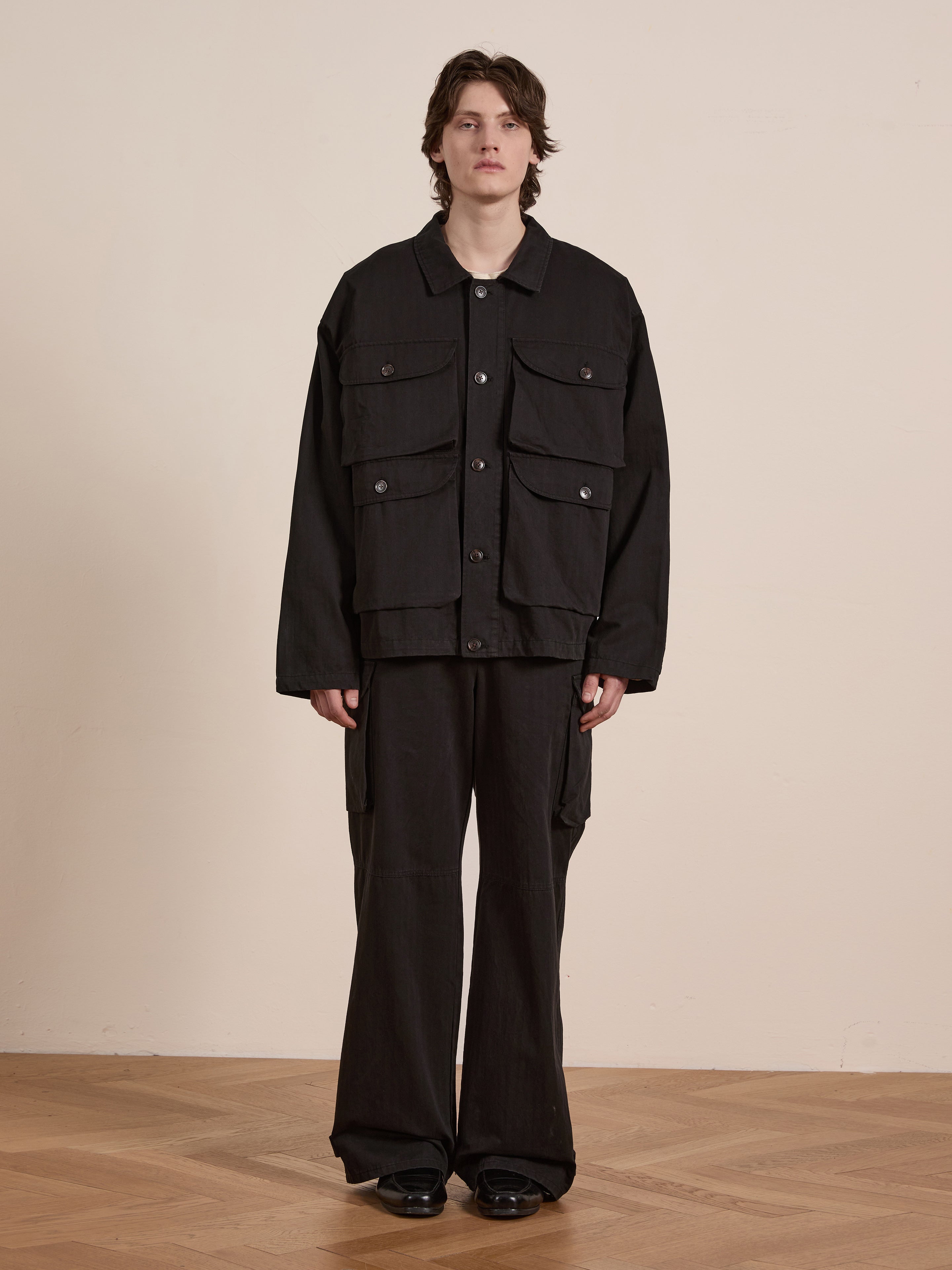 Wearing an oversized black Utility Herringbone Cargo Jacket and trousers from FOUND, a person stands on a wooden floor against a plain wall, exuding a vintage look.