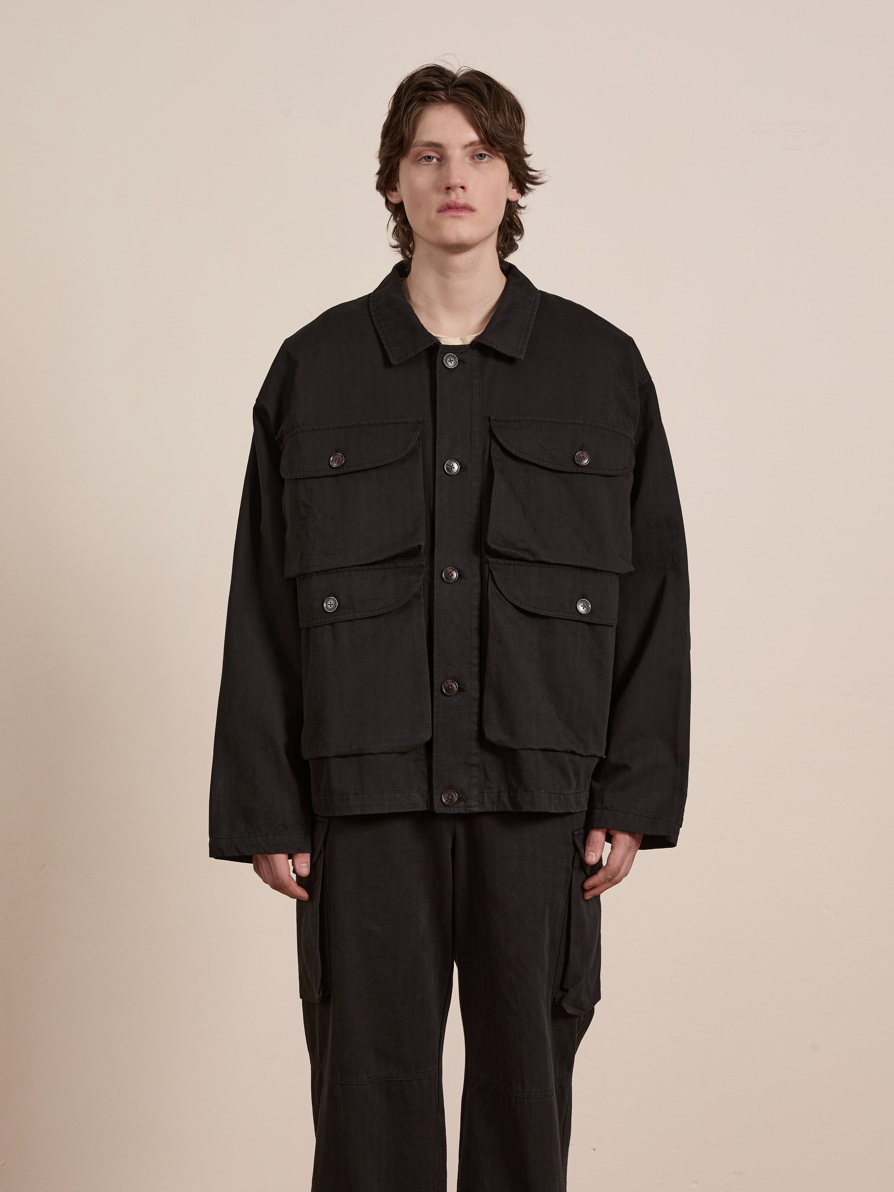 A person stands against a plain background, wearing a black Utility Herringbone Cargo Jacket by FOUND, featuring large pockets and a vintage look that seamlessly blends style and functionality.