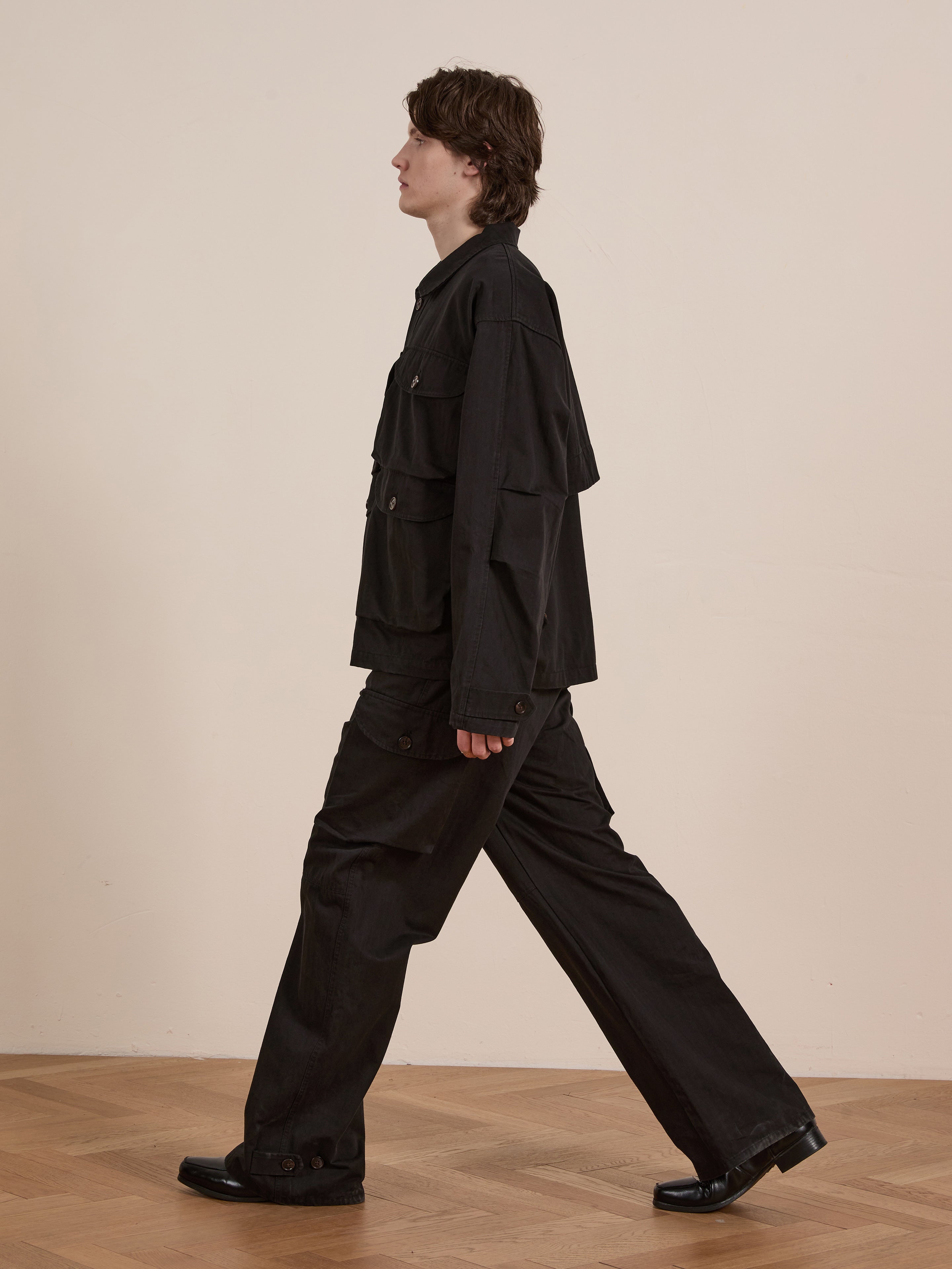 A person walks gracefully in profile across a wooden floor, dressed in a vintage-look black Utility Herringbone Cargo Jacket by FOUND and matching pants.