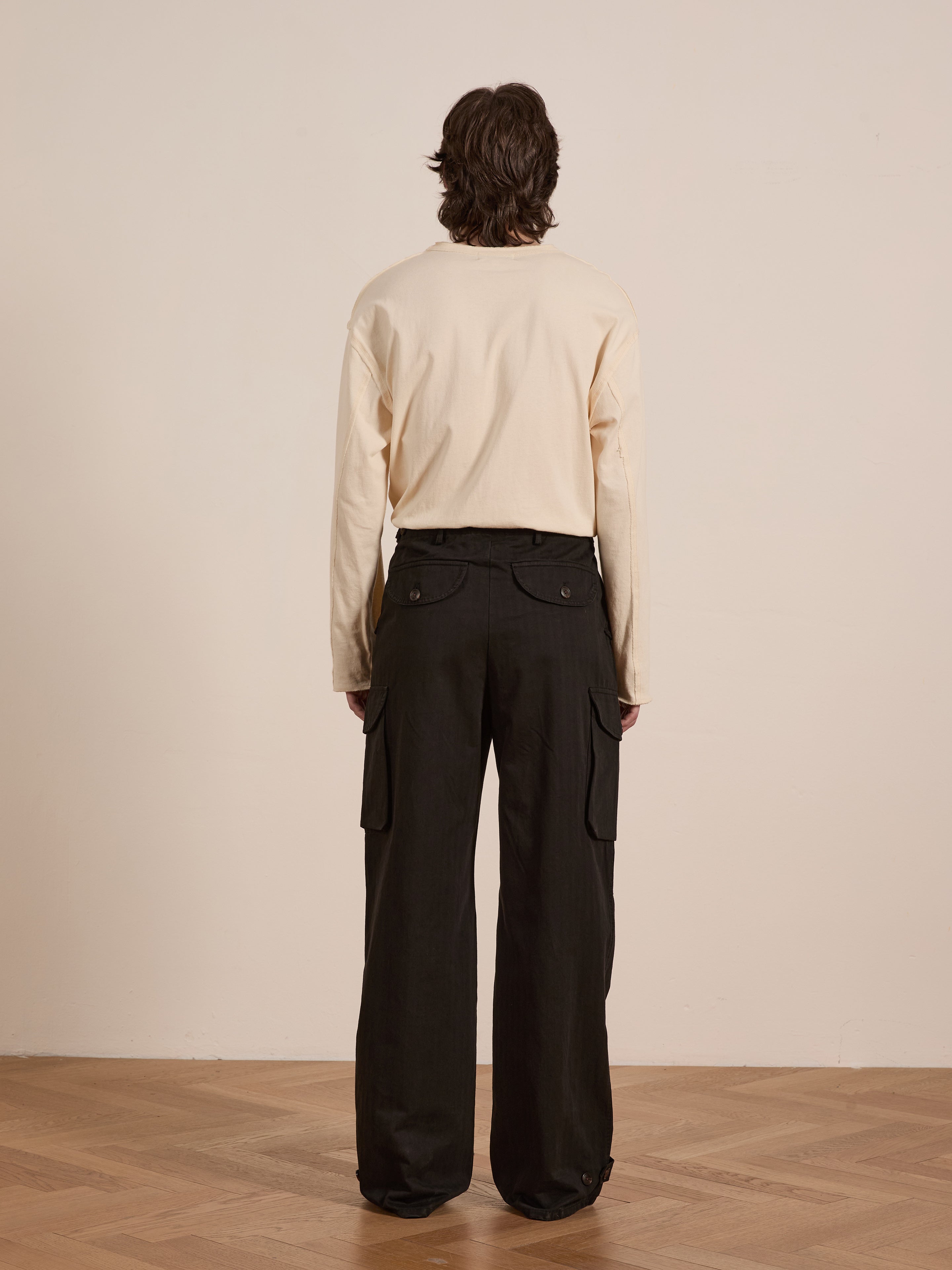 A person with long hair, in a cream long-sleeve shirt and FOUND's Utility Herringbone Cargo Pants, stands facing away on a wooden floor against a plain background.