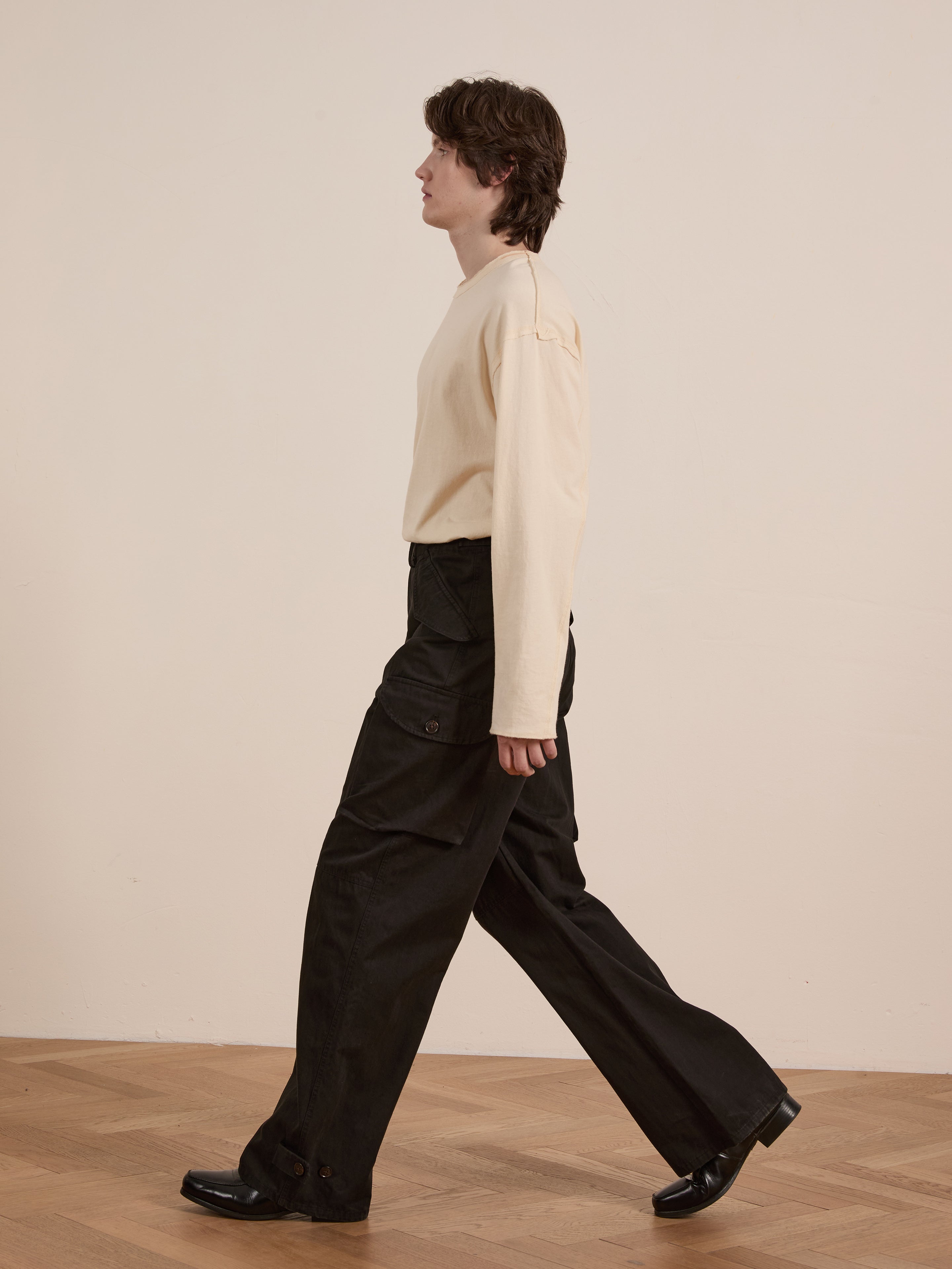 A person strolls indoors, donning a beige long-sleeve shirt and FOUND's Utility Herringbone Cargo Pants, which offer a vintage flair. Paired with black shoes, they tread confidently across the wooden floor.