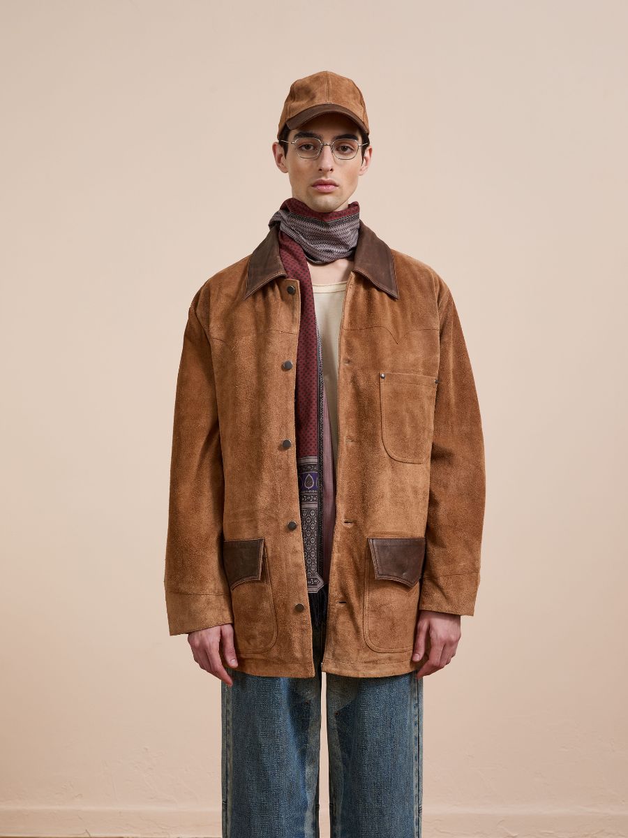 A person faces forward against a beige background, wearing the FOUND Suede Contrast Chore Jacket in a unisex relaxed fit, paired with a brown cap, glasses, patterned scarf, light shirt, and blue jeans.