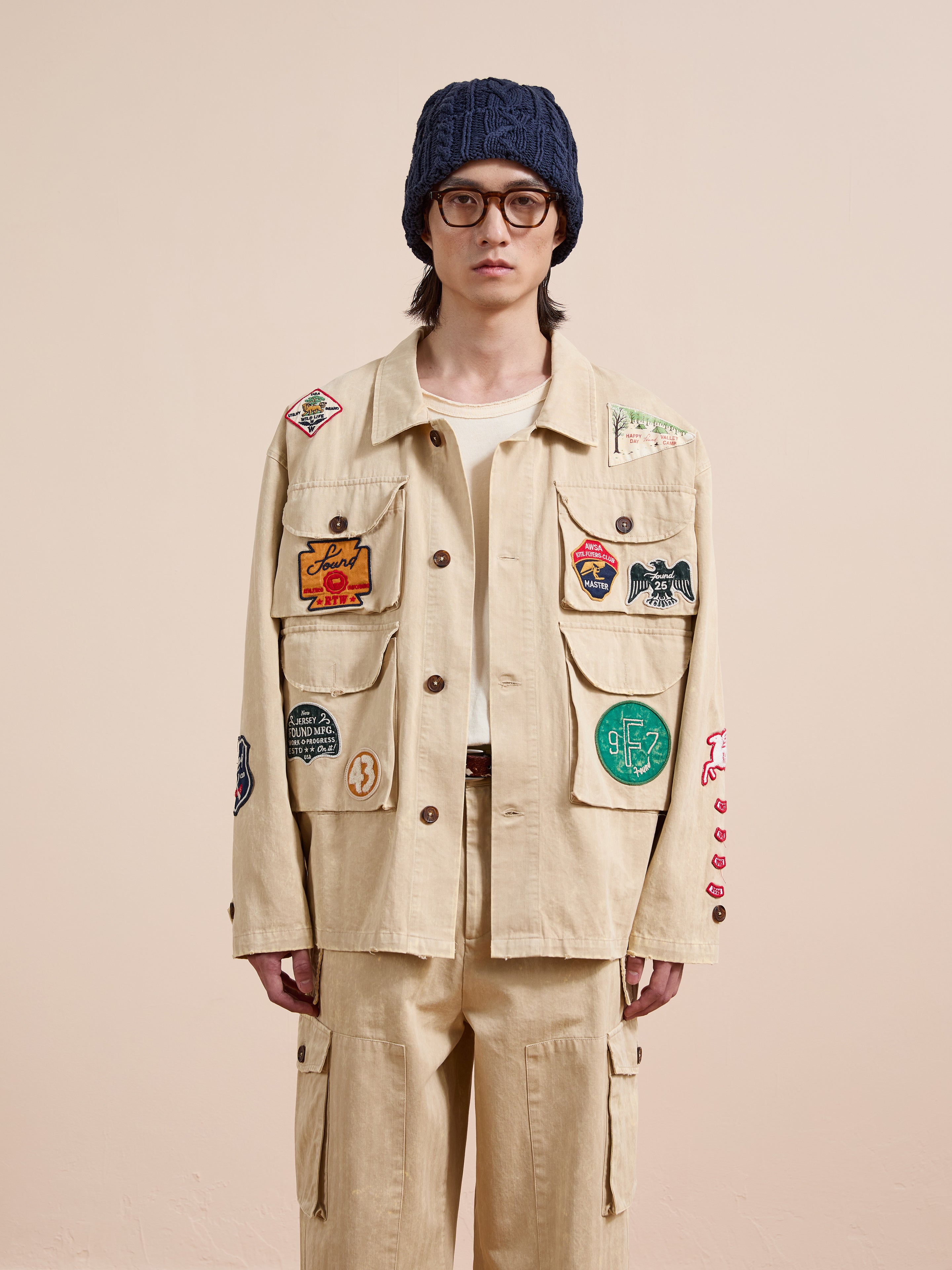 A person wearing the FOUND Herringbone Utility Patchwork Jacket with matching pants, a white shirt, round glasses, and a dark blue knit beanie, stands against a plain background.
