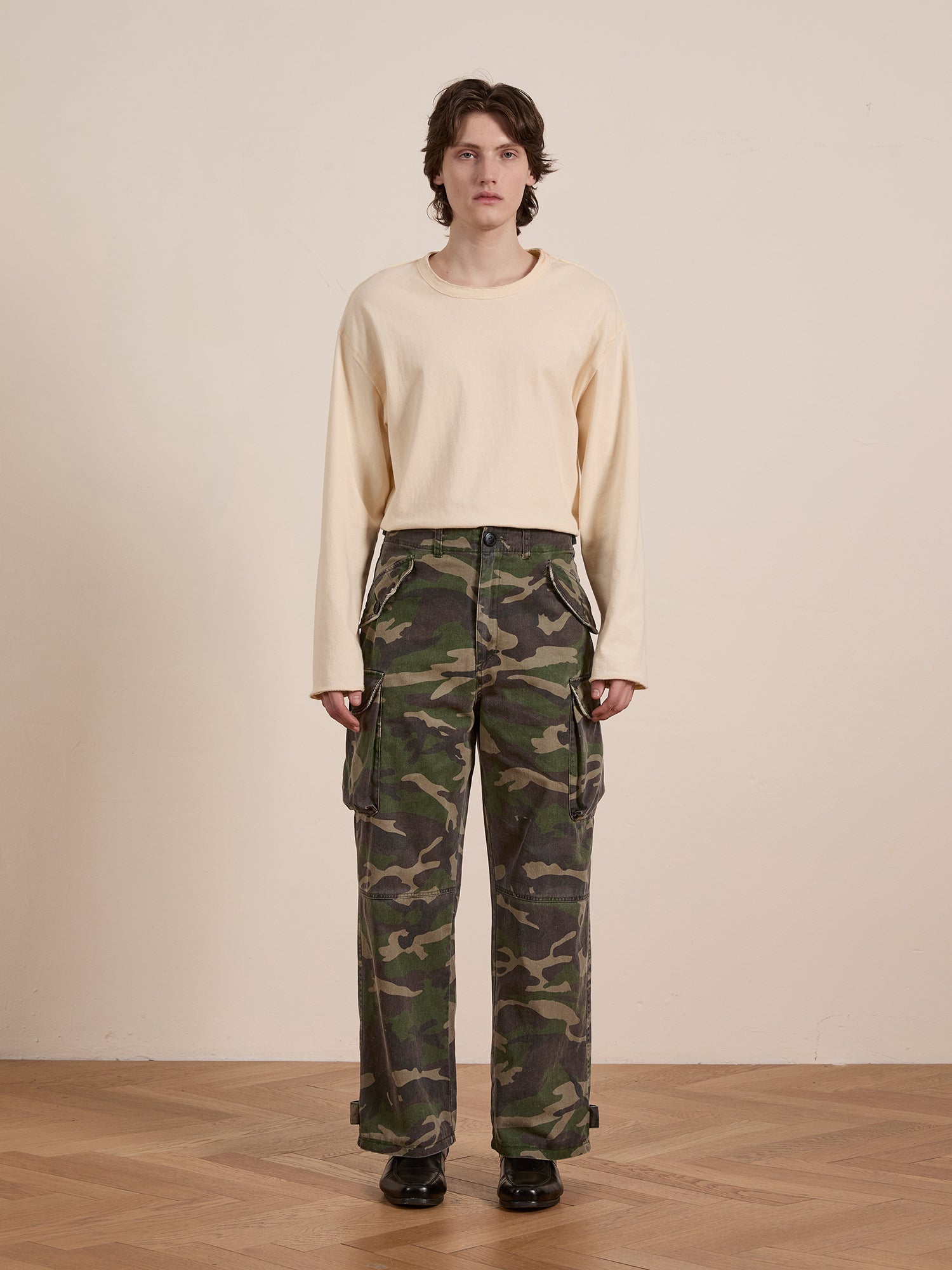 Indoors, a person flaunts a vintage style with a beige long-sleeve shirt and FOUND's Camouflage Twill Utility Cargo Pants. Their short hair enhances the look, while black shoes offer bold contrast against the simple backdrop.