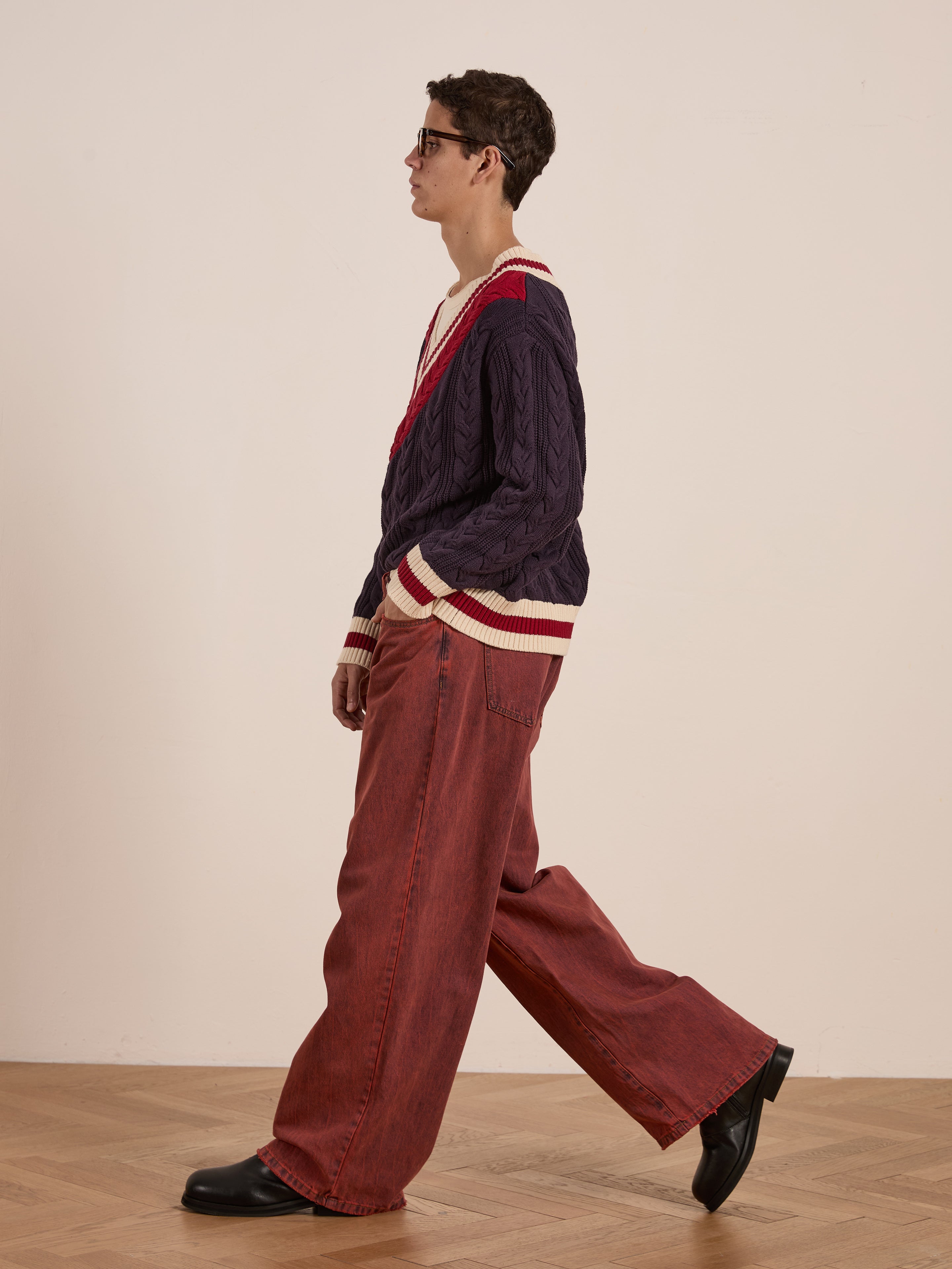 A person in a purple FOUND V-Neck Cricket Sweater with red and white trim, red wide-leg pants, and black shoes walks on a wooden floor.