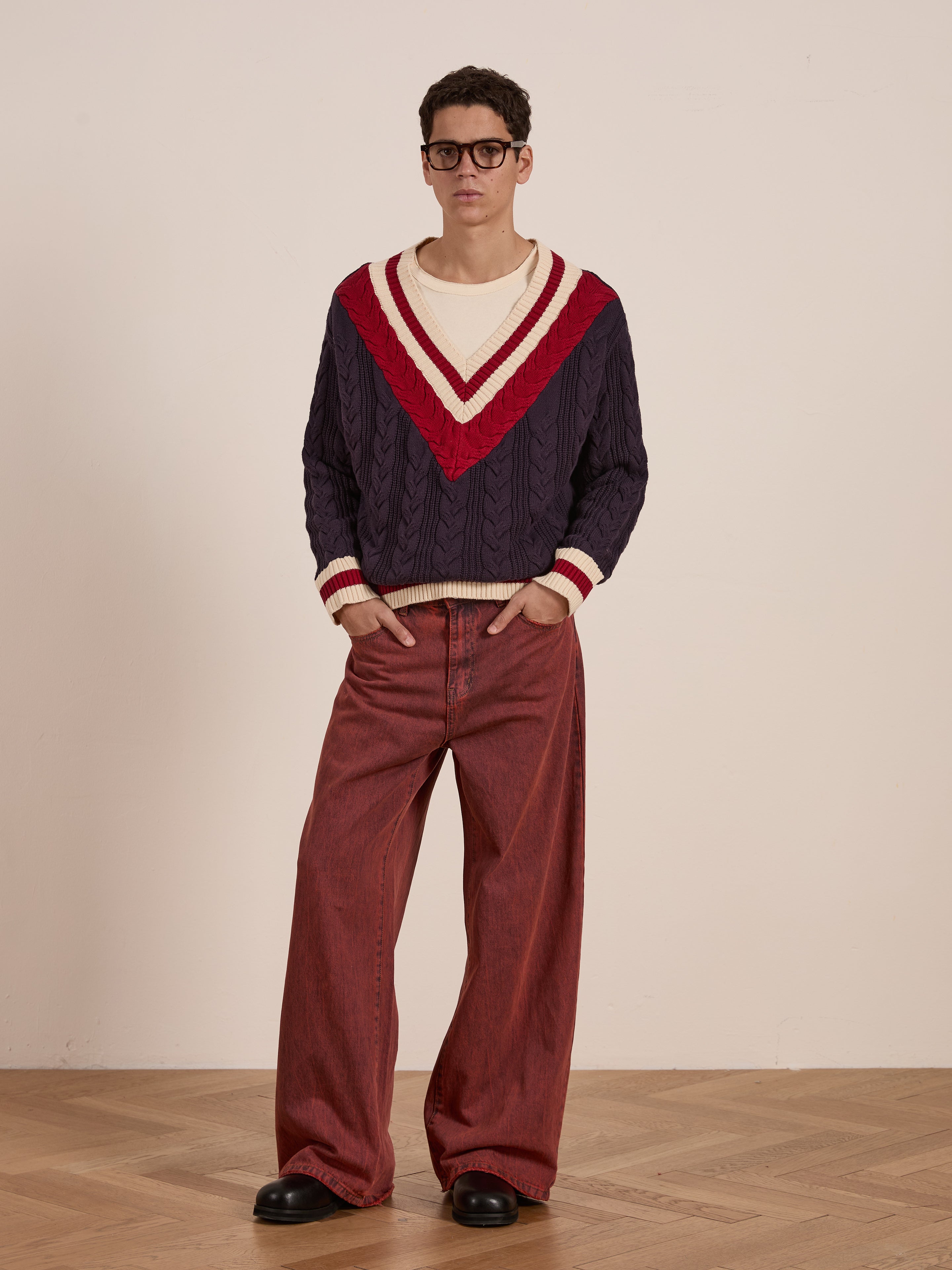 A person wears the FOUND V-Neck Cricket Sweater in navy with cream accents, paired with red wide-leg pants and black shoes, standing on a wooden floor against a plain backdrop.