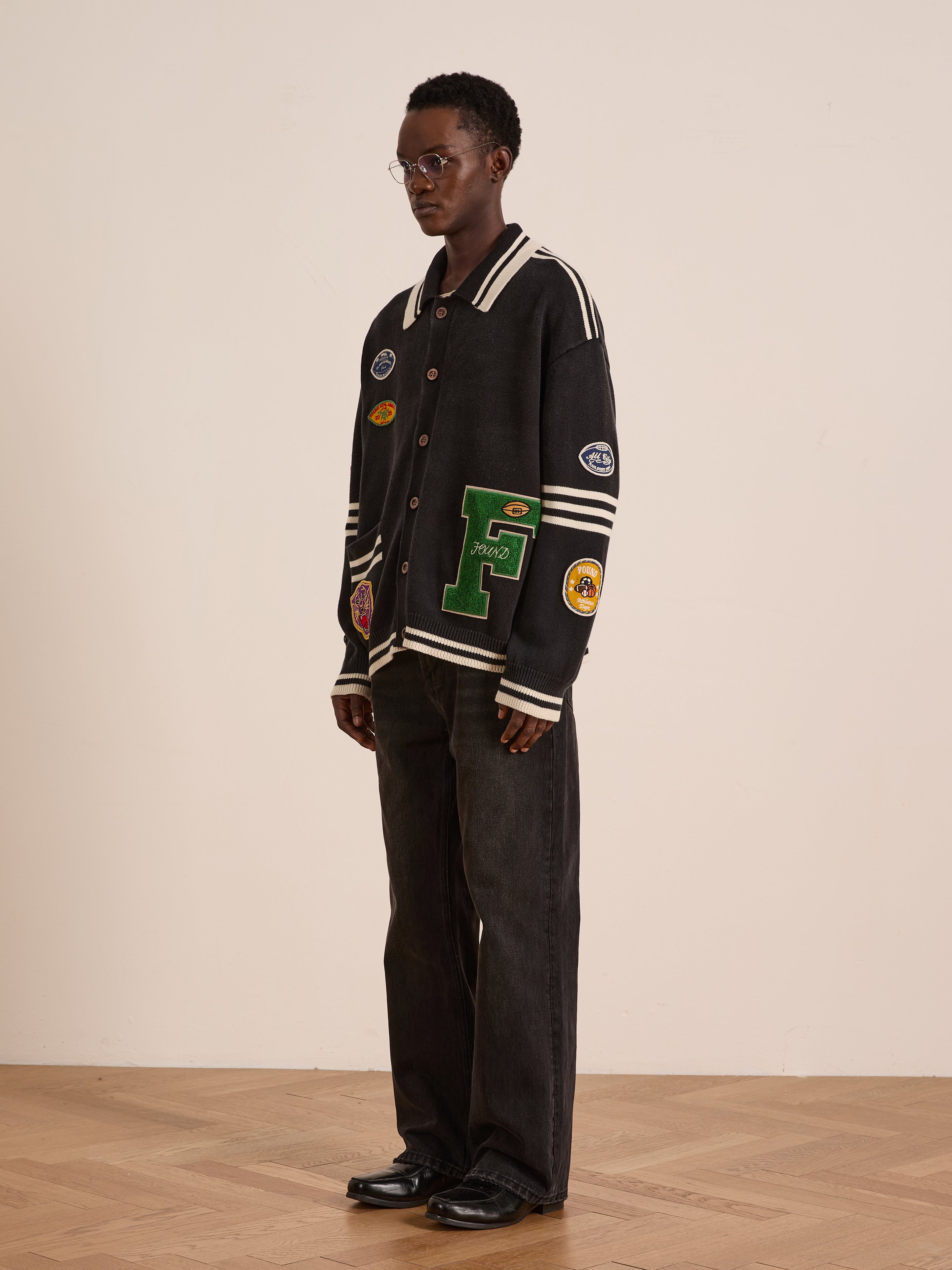 A person stands indoors against a plain background wearing FOUND’s Varsity Patch Collared Cardigan, black jeans, black shoes, and glasses.