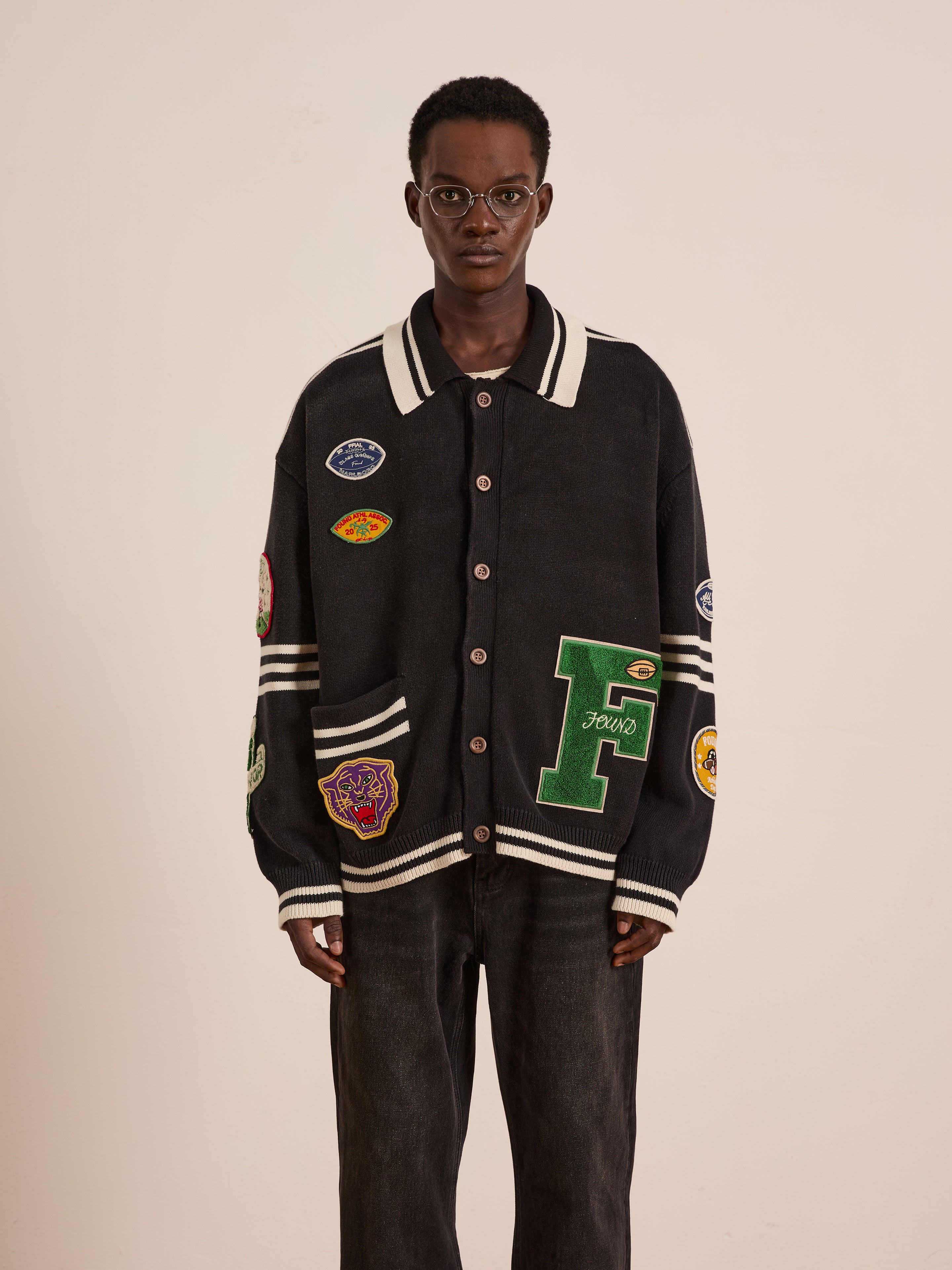 A person wearing glasses, the FOUND Varsity Patch Collared Cardigan, and black pants stands facing forward against a plain light background.