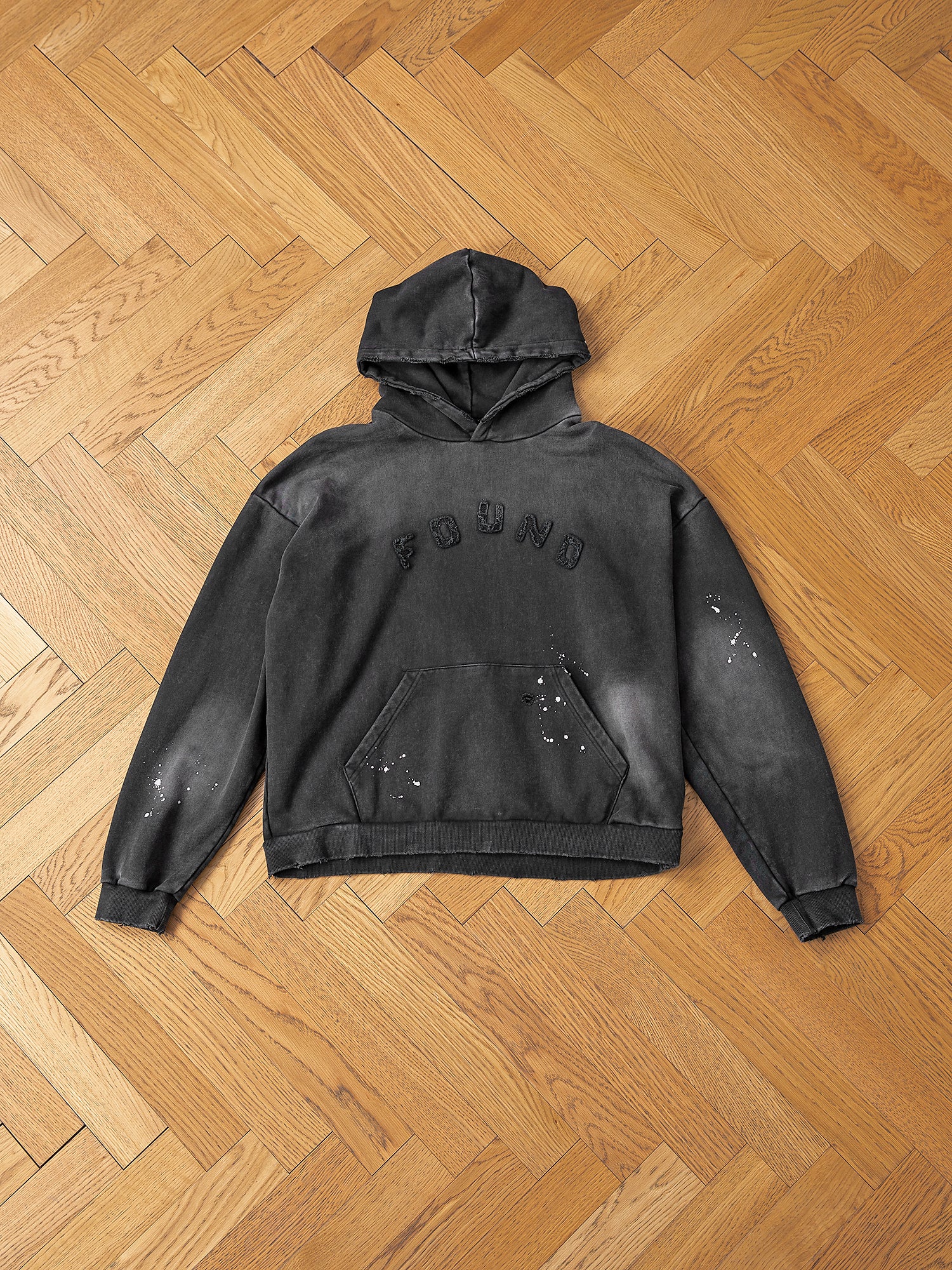 A "FOUND" Logo Patch Hoodie in vintage black, crafted from soft French terry cotton with a slightly distressed look, lying flat on a wooden herringbone floor.