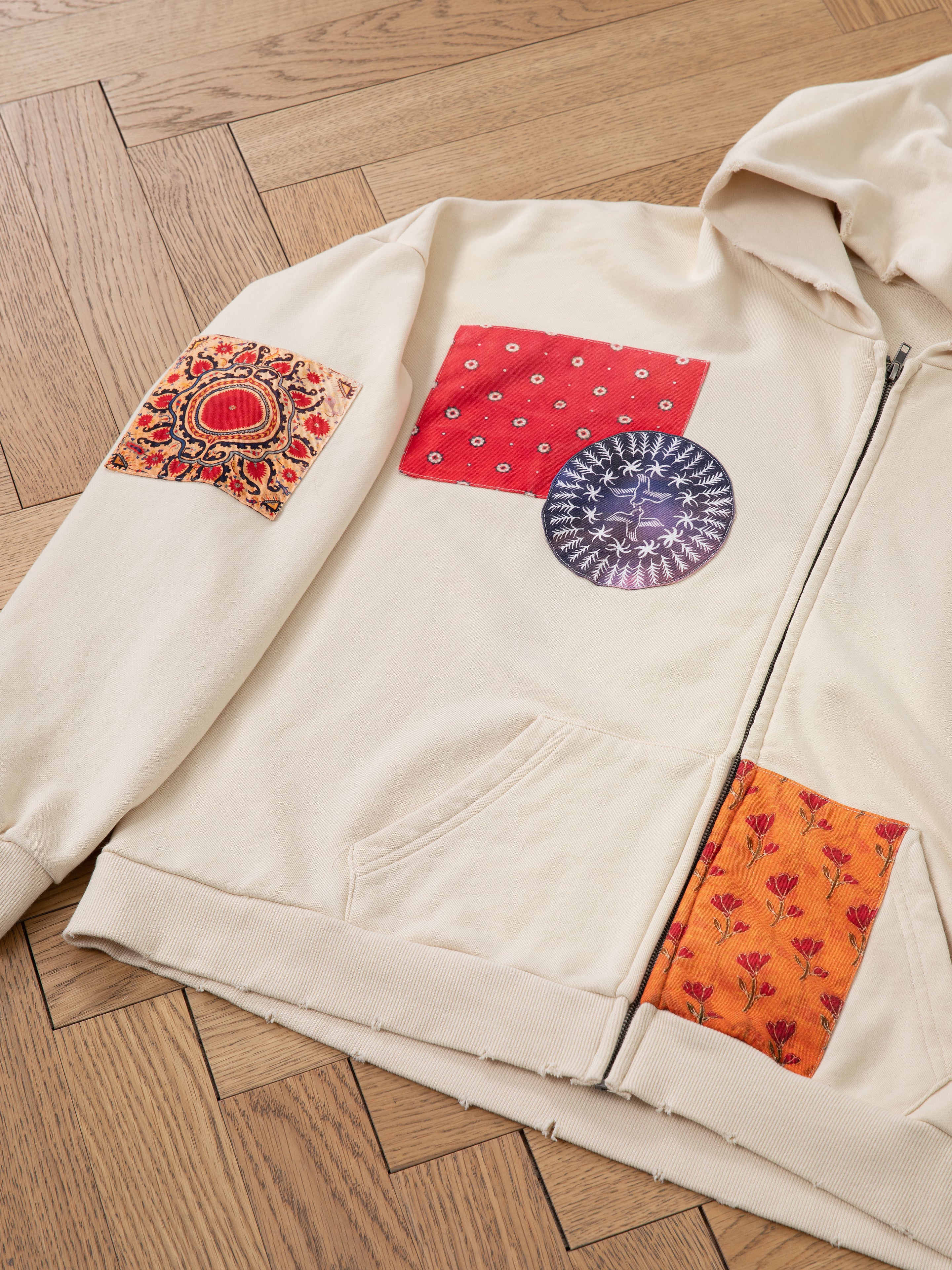 A Tapestry Patchwork Zip Up Hoodie by FOUND, featuring a cream design in beige French terry cotton, embellished with four colorful fabric patches inspired by South Asian tapestry patterns in circular and square shapes, is laid out on a wooden herringbone floor.