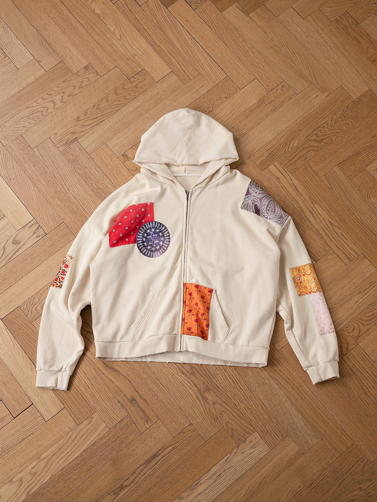 A Tapestry Patchwork Zip Up Hoodie from FOUND, showcasing South Asian tapestry patterns in vibrant red, purple, and orange, lies on a wooden floor.